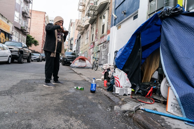 A person stands near a tent on the sidewalk of a street while they eat a bowl of soup. The street is lined with cars and surrounded housing buildings.