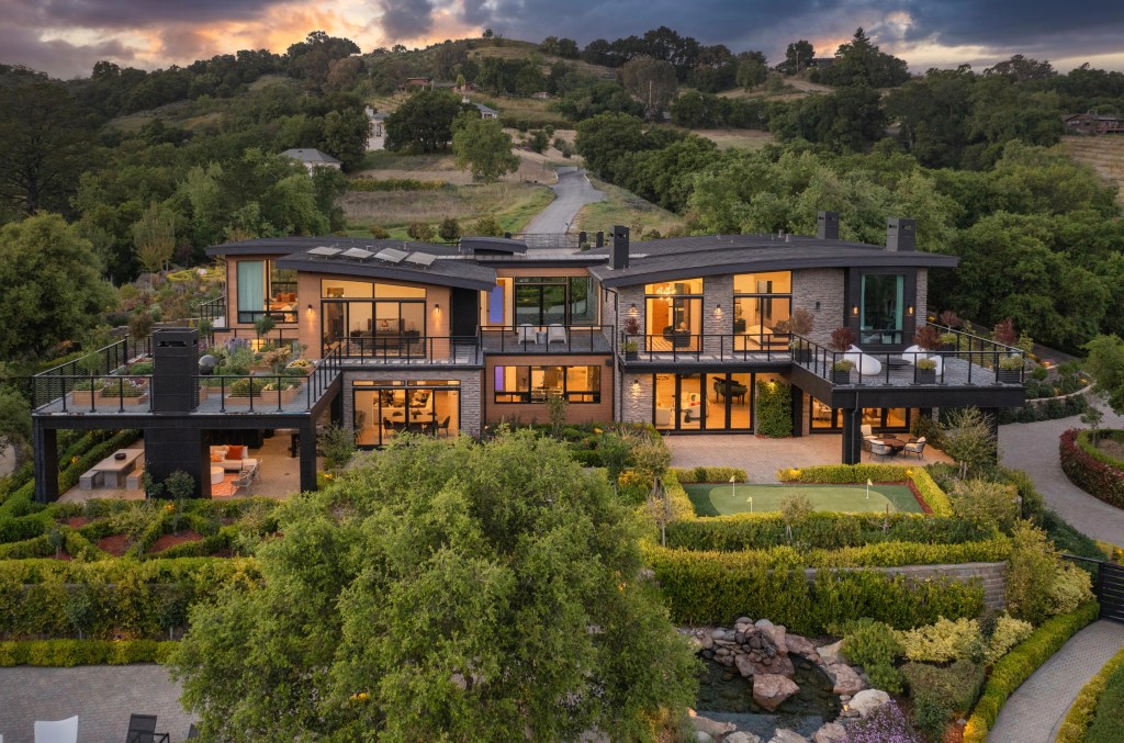 An aerial view of a luxurious modern home with multiple balconies and terraces surrounded by lush green hills, featuring a putting green and outdoor dining areas.