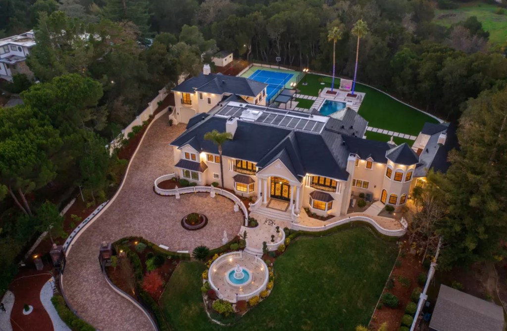 Aerial view of a large estate home with a circular driveway, fountain, pool, and tennis court, surrounded by trees.