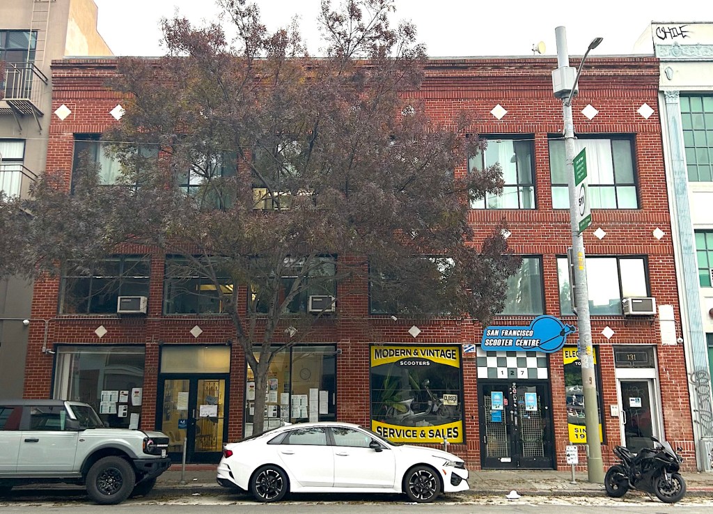 A side shot of a brick building with a tree and cars parked in front.