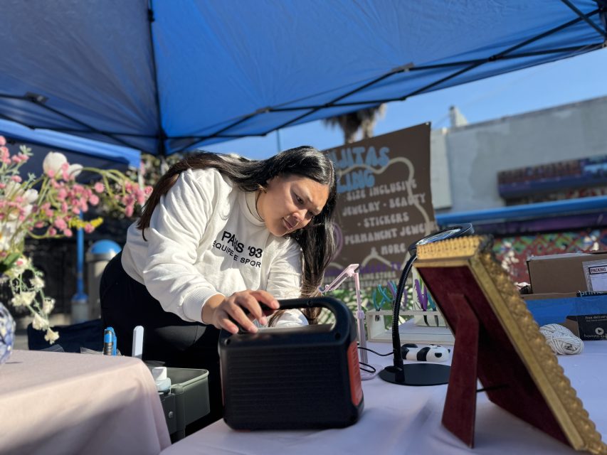 A woman adjusts equipment on a table under a blue canopy at an outdoor event, with flowers and framed items nearby.