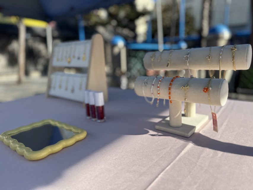 A display of bracelets, necklaces, and lip glosses on a table with a small mirror at an outdoor jewelry stand.