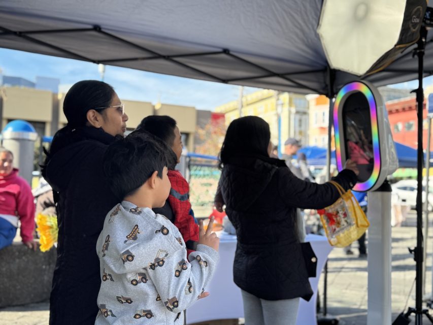 A woman and two children stand near a photo booth while another person interacts with the booth under a canopy at an outdoor event.