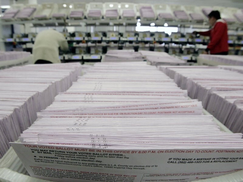 Ballots are stacked in boxes. There are people in the background inspecting some.