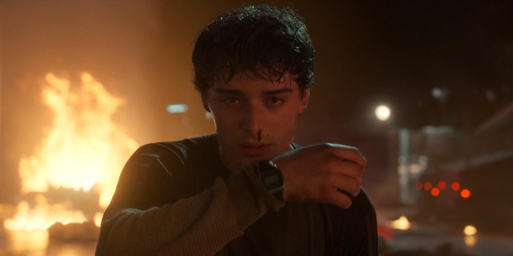 A young man with a bloody nose stares ahead as a flames engulf the area behind him
