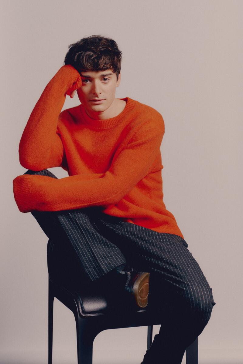 A young man wearing jeans and an orange sweater poses on a chair