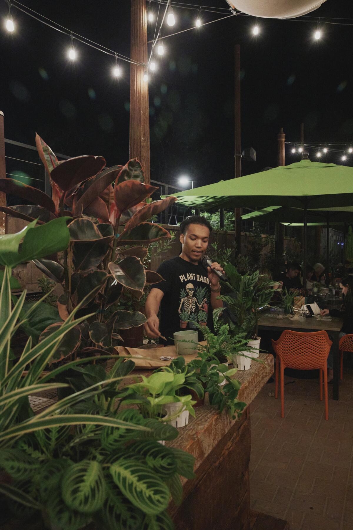 A man standing by potted plants calls out plant names on a microphone. 