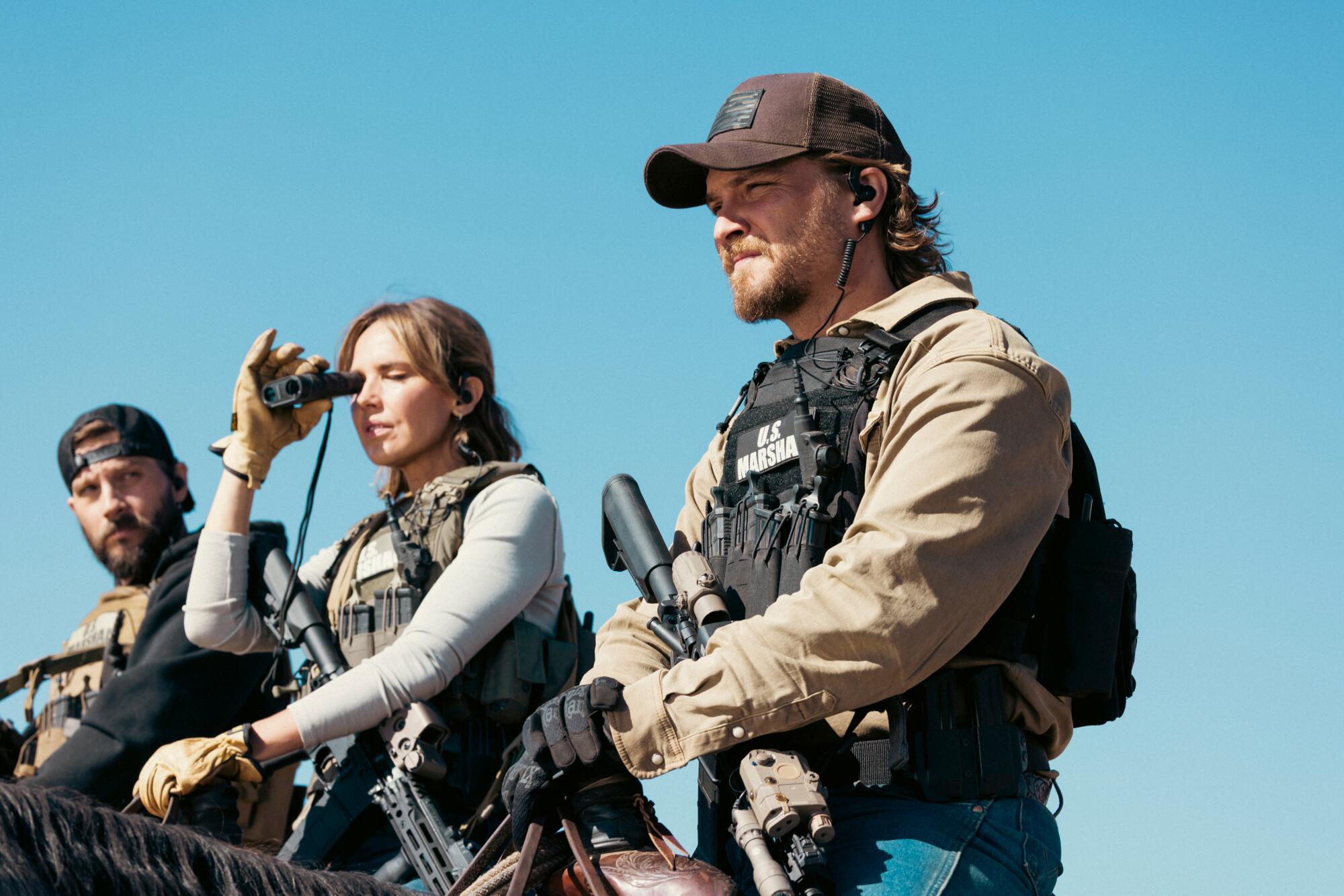 A woman holding up a pair of binoculars is flanked by two men on horseback.