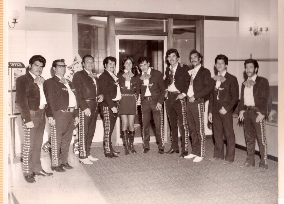 Ten people stand in a line facing the camera in a black and white photo. They wear traditional mariachi dark jackets and boots.