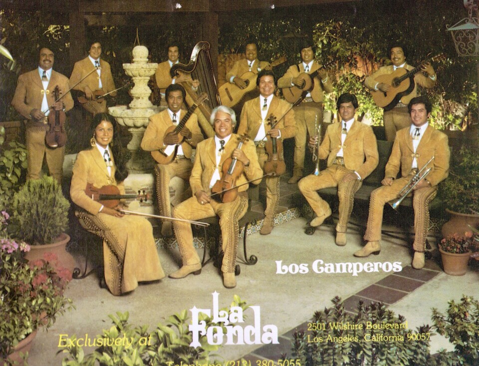 Twelve people hold musical instruments. They wear the same, mustard-colored outfits. One of them a woman, wears a long skirt.