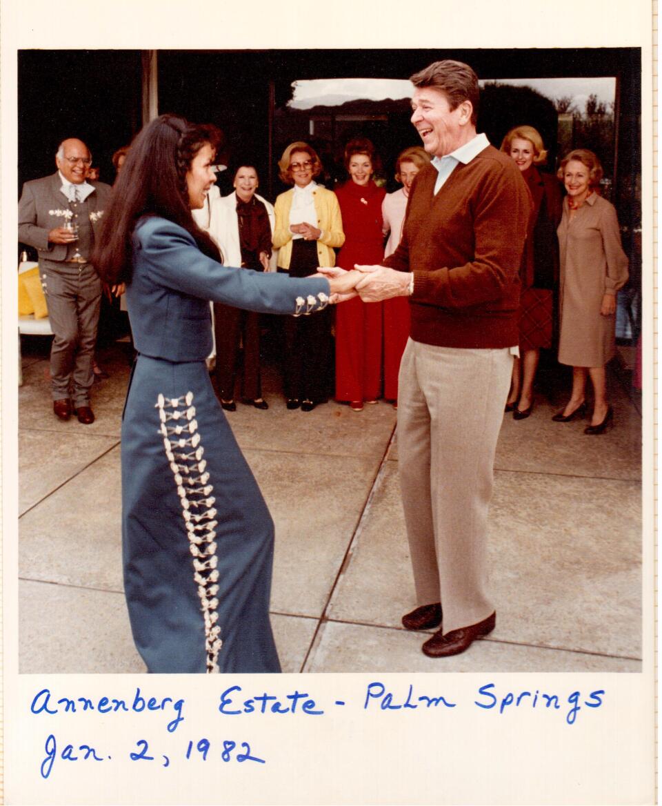 A woman with long dark hair and medium skin, wearing a blue jacket and long skirt, is dancing with a light skinned man wearing a maroon v neck sweater and grey pants.