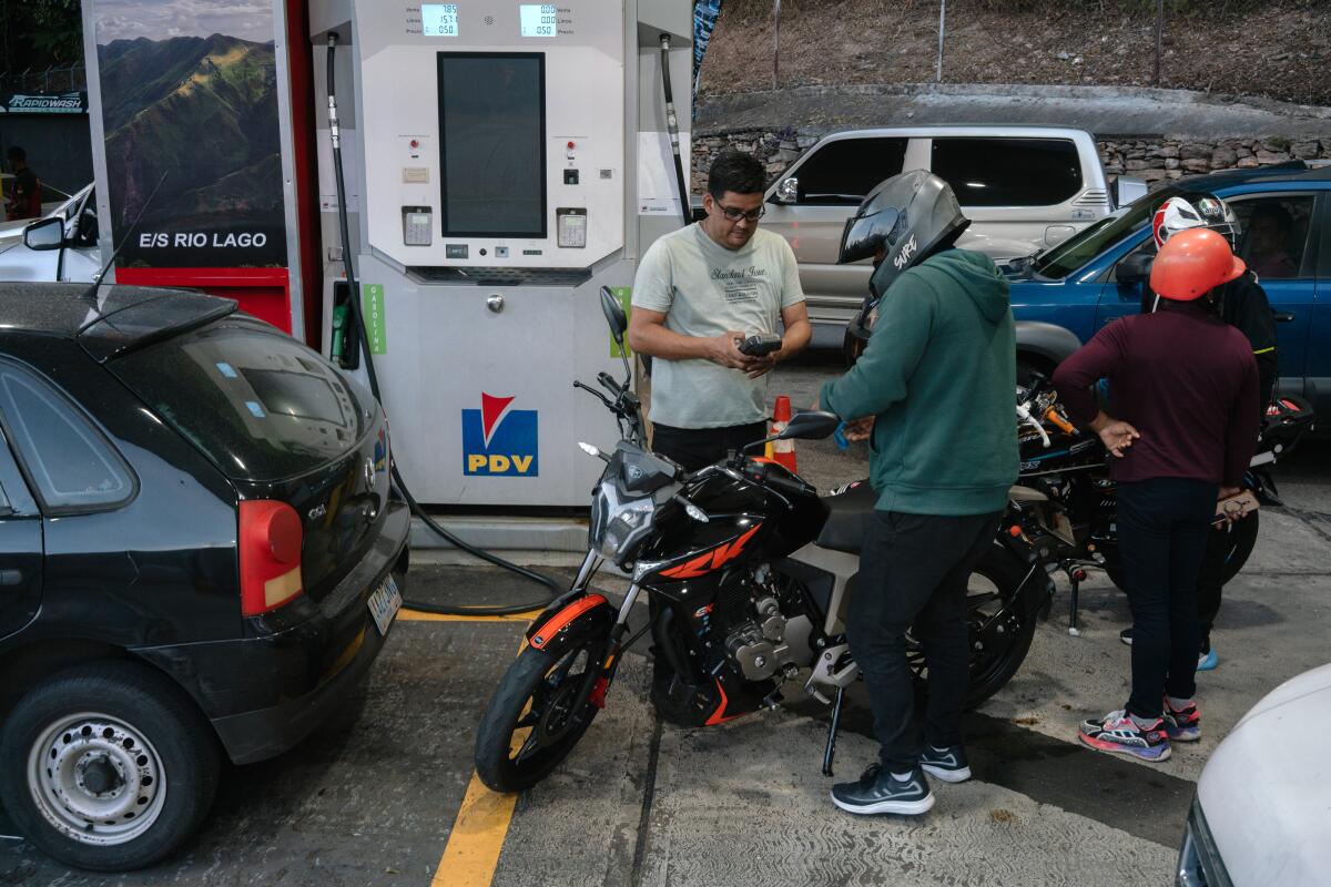 People stand around cars and a motorbike at a crowded gas pump.