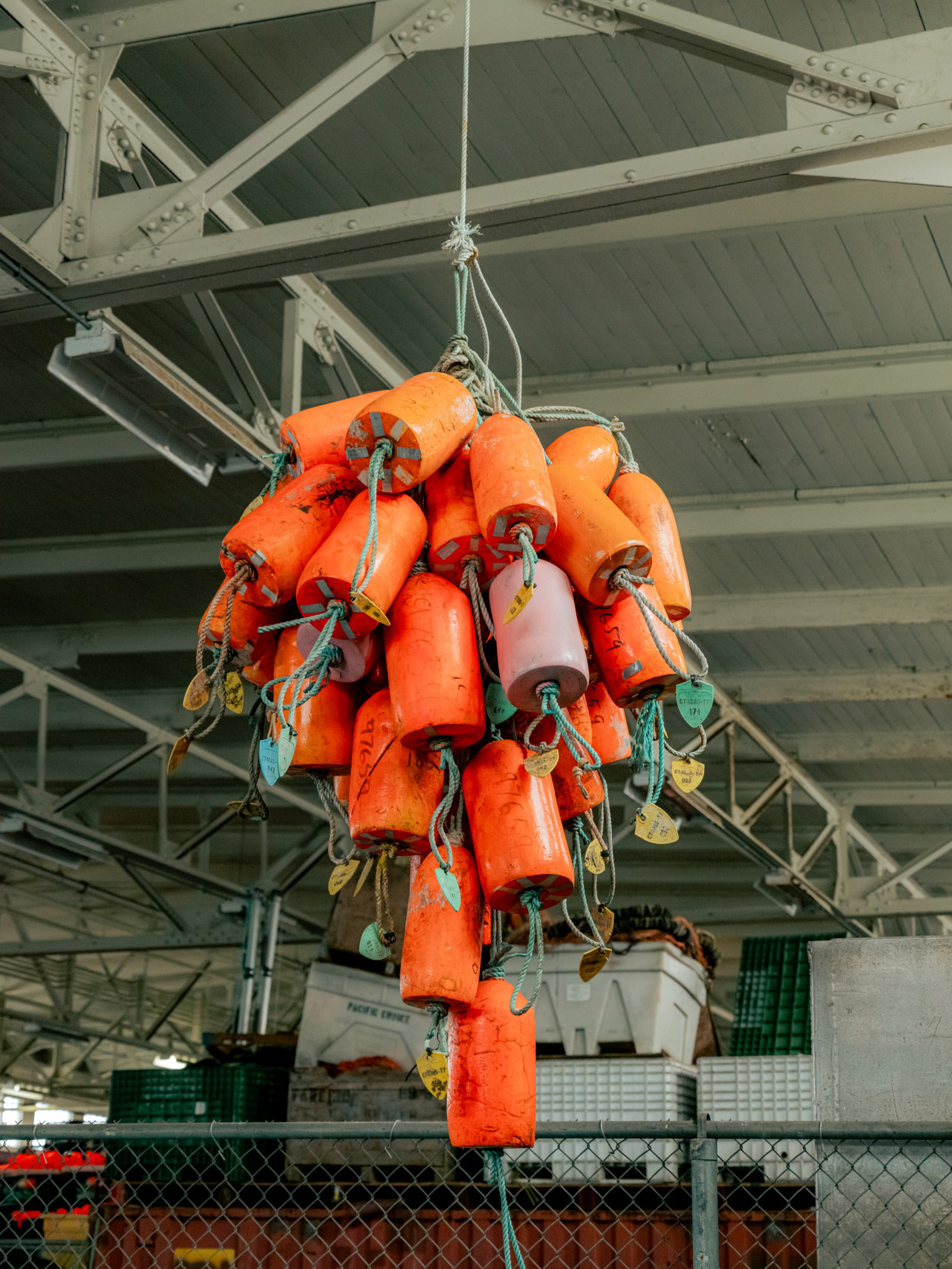 A cluster of worn orange buoys with attached ropes hangs from the ceiling inside a storage or market space.