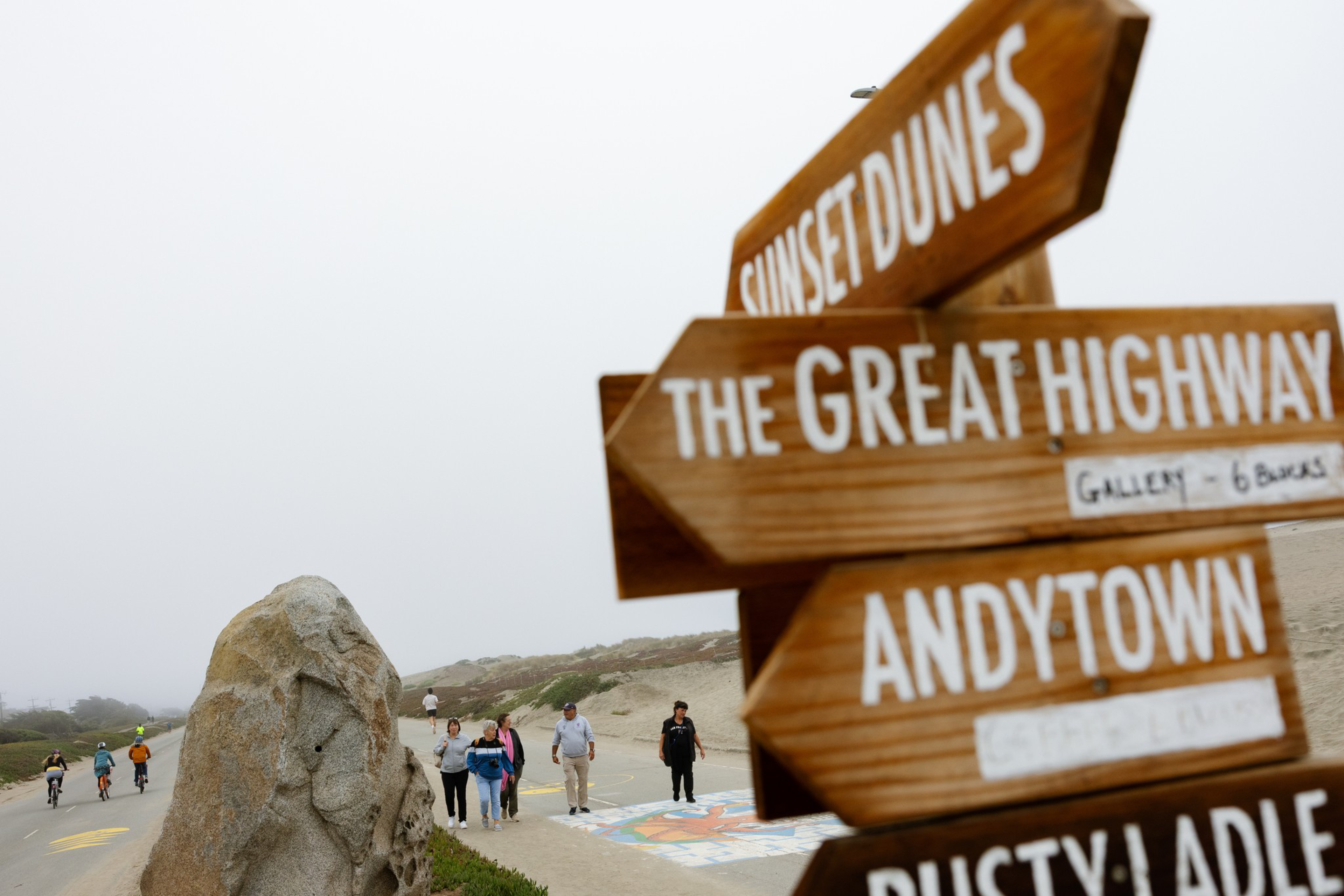 Wooden signs point towards Sunset Dunes, The Great Highway, and Andytown near a beach path where people walk beside a large rock and sandy dunes.