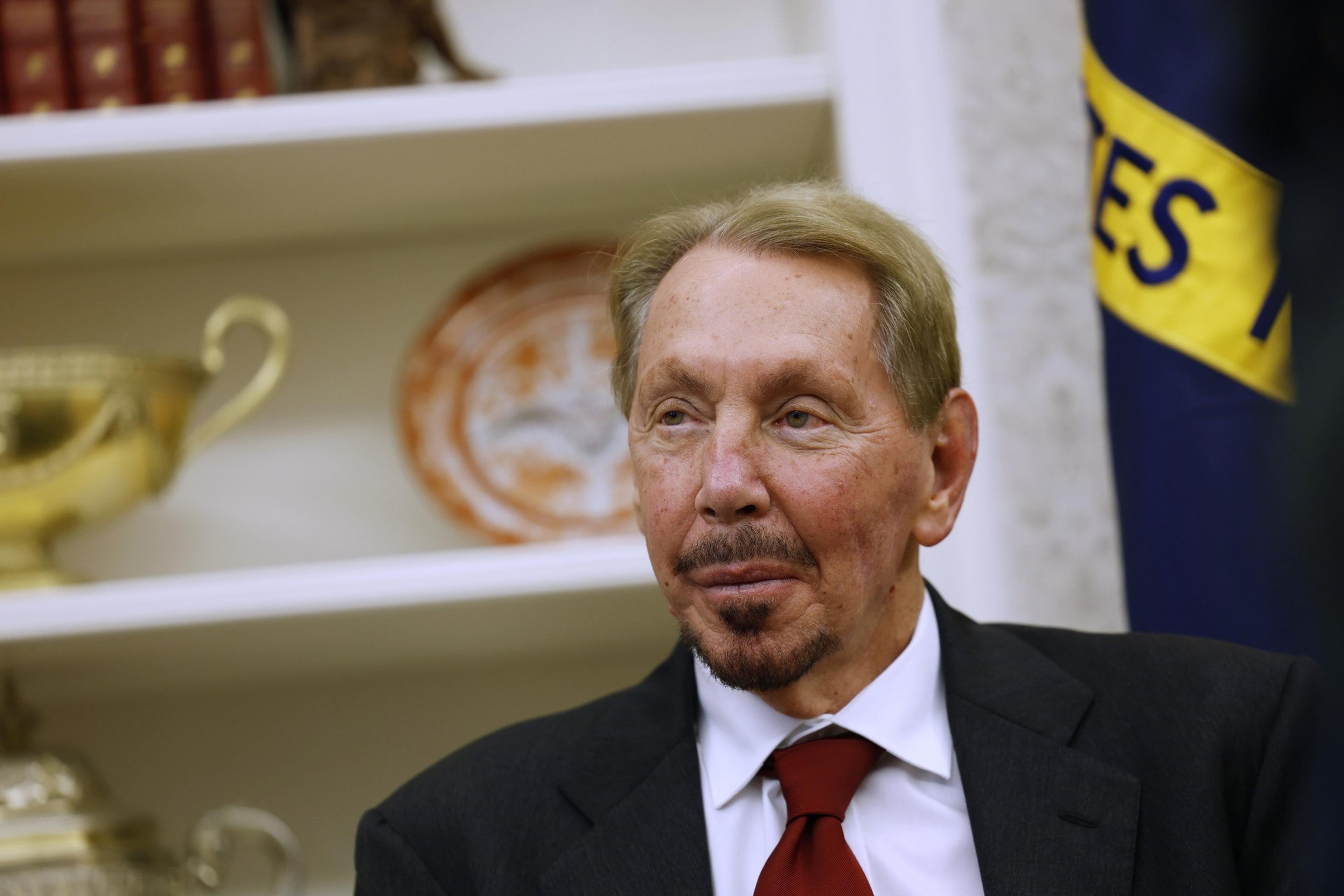 A man with light brown hair and a mustache wearing a dark suit and red tie sits in front of shelves with decorative items.
