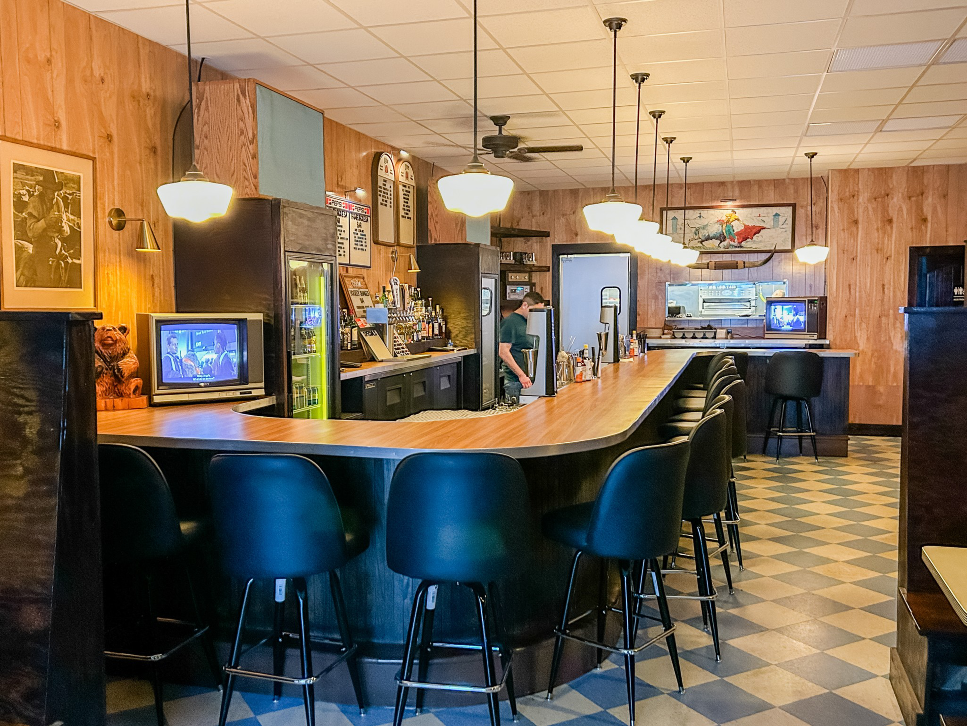A retro-style bar with a wood-paneled interior, high black stools, hanging lights, two small TVs, and a person behind the counter.