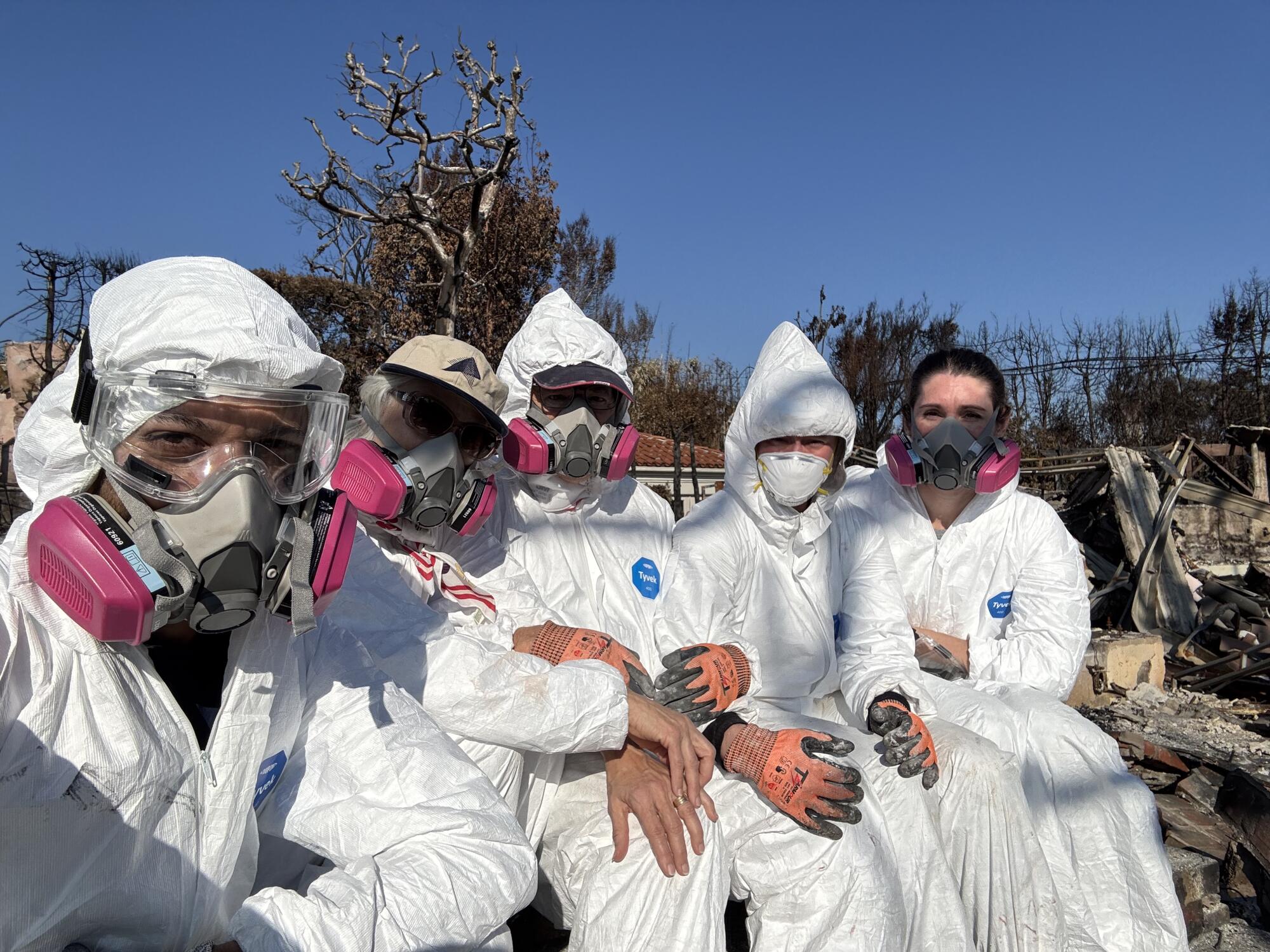 Five people in white hazmat suits and respirators sitting on the burned rubble of a home.
