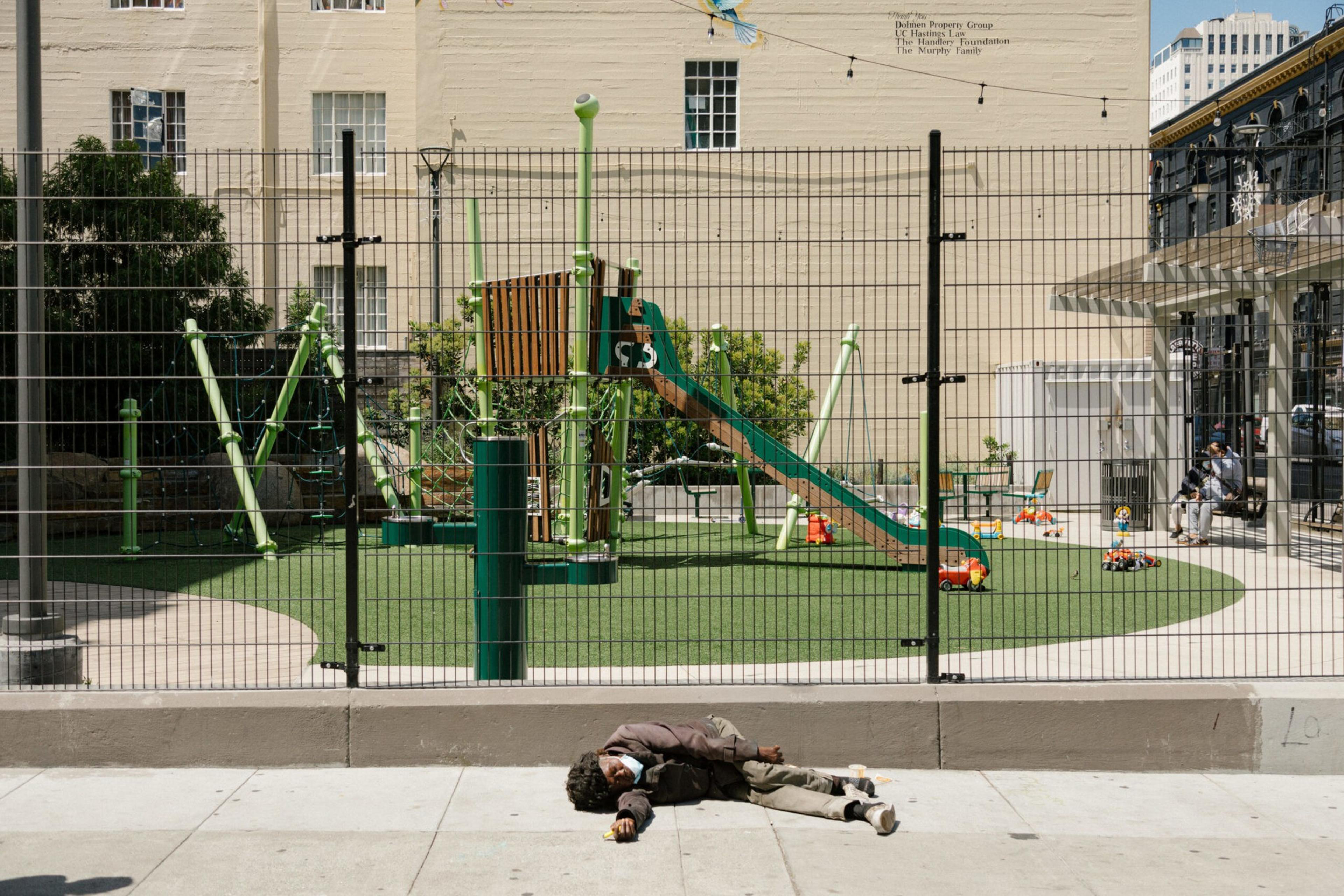 A man lies on the sidewalk in front of a fenced playground with green play structures. Inside, two people sit on a bench, and colorful toys are scattered around.