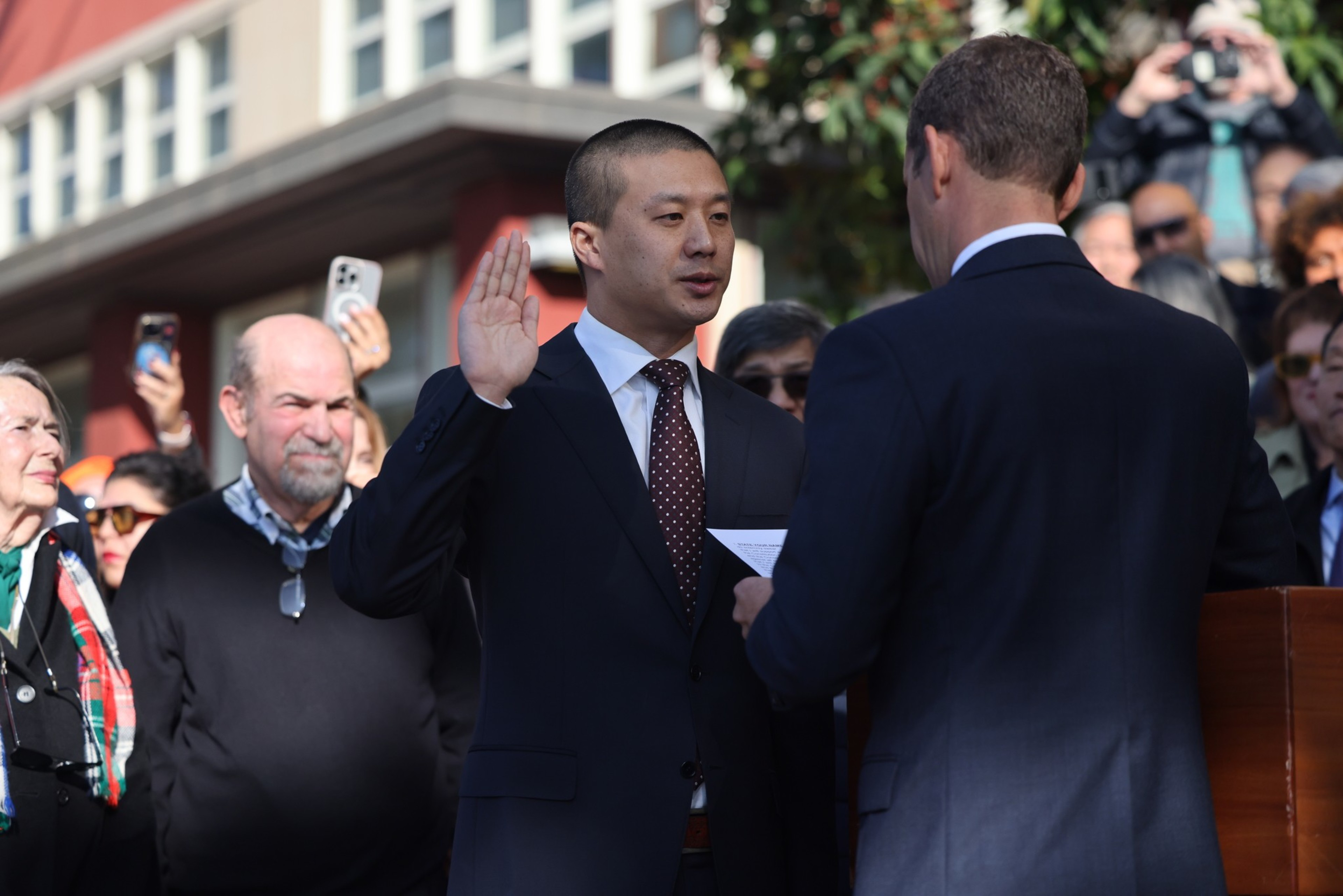 A man in a suit raises his right hand, taking an oath from another man holding a document, while a crowd watches and records the event.