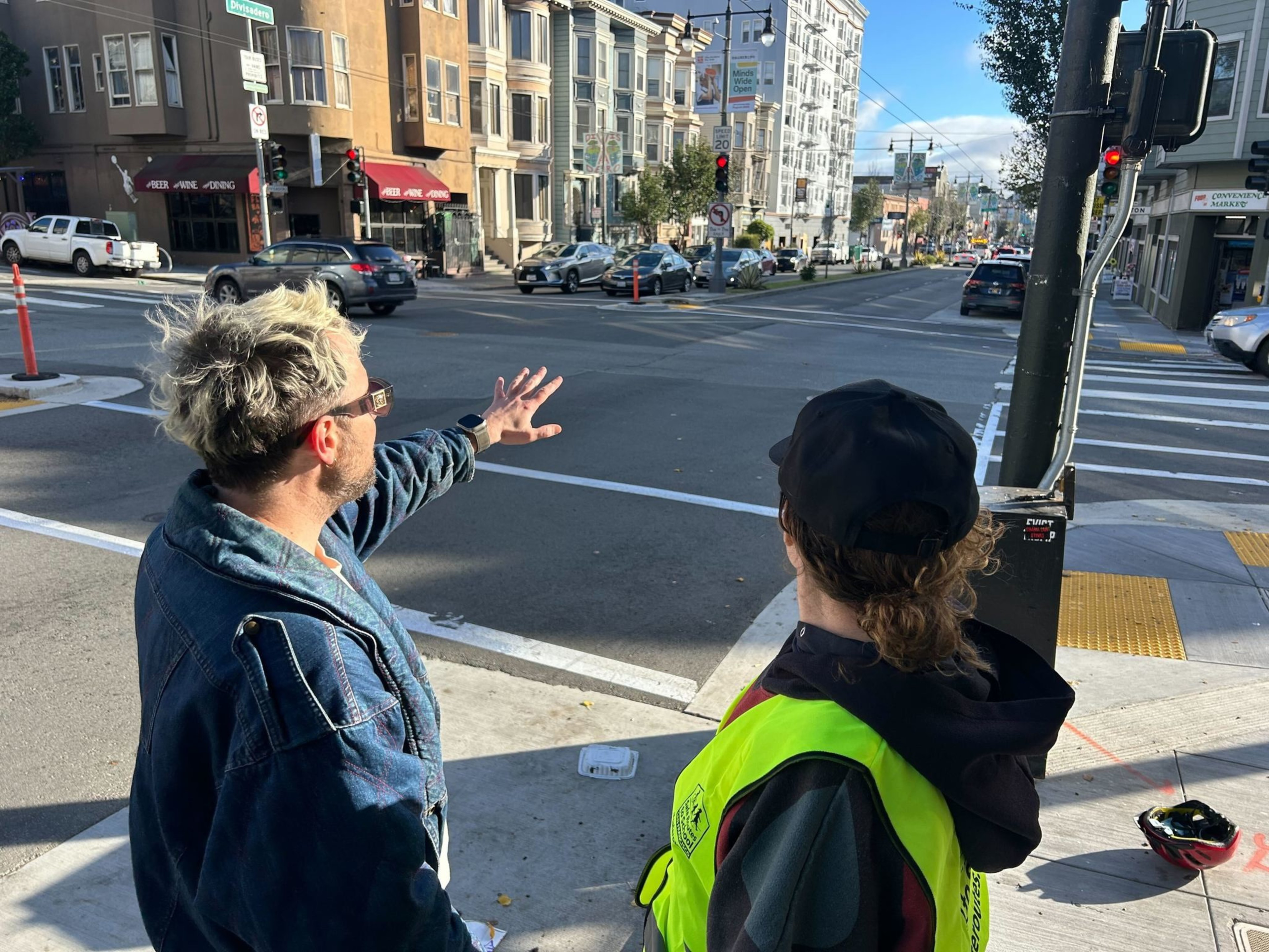 Two people stand on a sidewalk at an intersection; one person gestures across the street while the other wears a safety vest and black cap.
