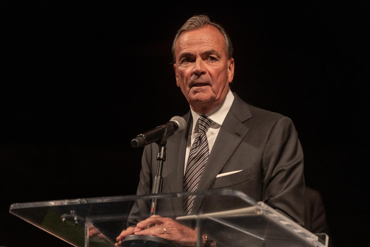 Rick Caruso stands in a suit at a lectern against a black background