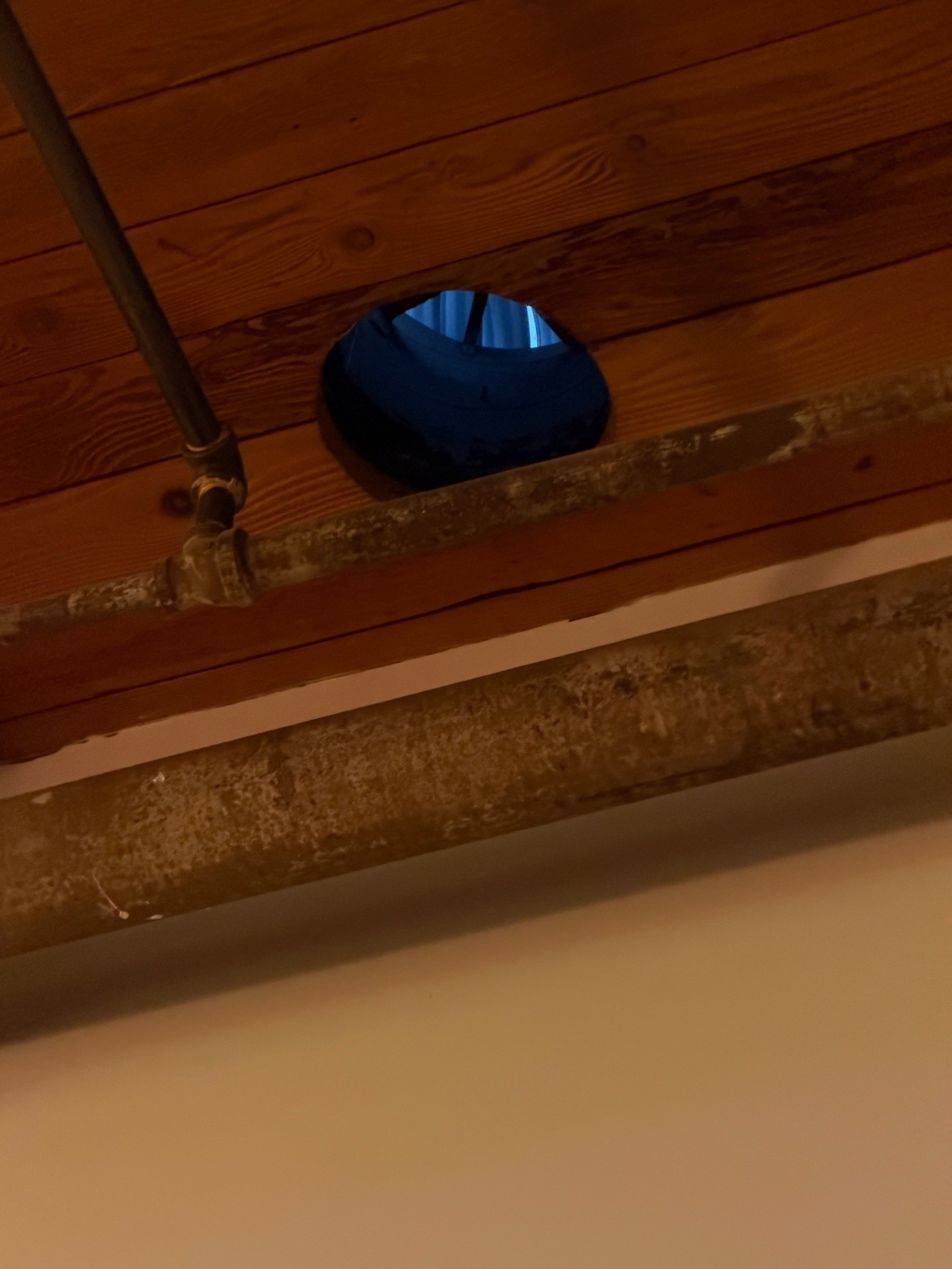 A wooden ceiling beam with a circular hole reveals a blue pipe inside, above two rusty metal pipes running horizontally.