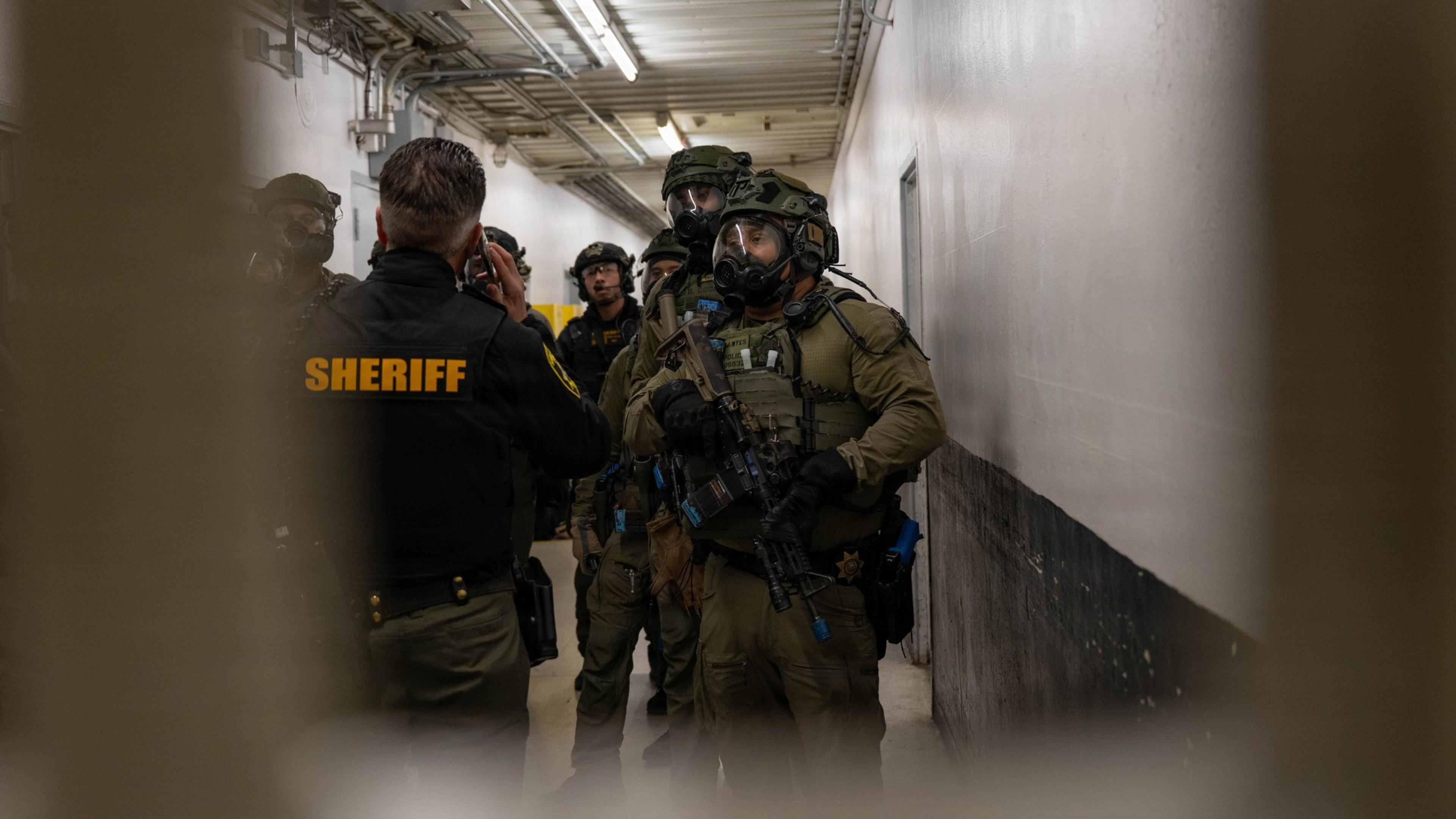 A group of armed tactical officers in full gear and helmets stand in a narrow hallway, facing a sheriff in a black jacket with “SHERIFF” in yellow letters.