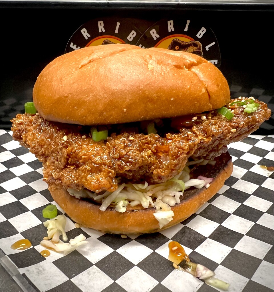 A fried chicken sandwich on a toasted brioche bun features a large crispy chicken cutlet coated in orange glaze and sesame seeds, topped with shredded cabbage, scallions, and sauce, served on black and white checkered paper with the Terrible Burger logo in the background.