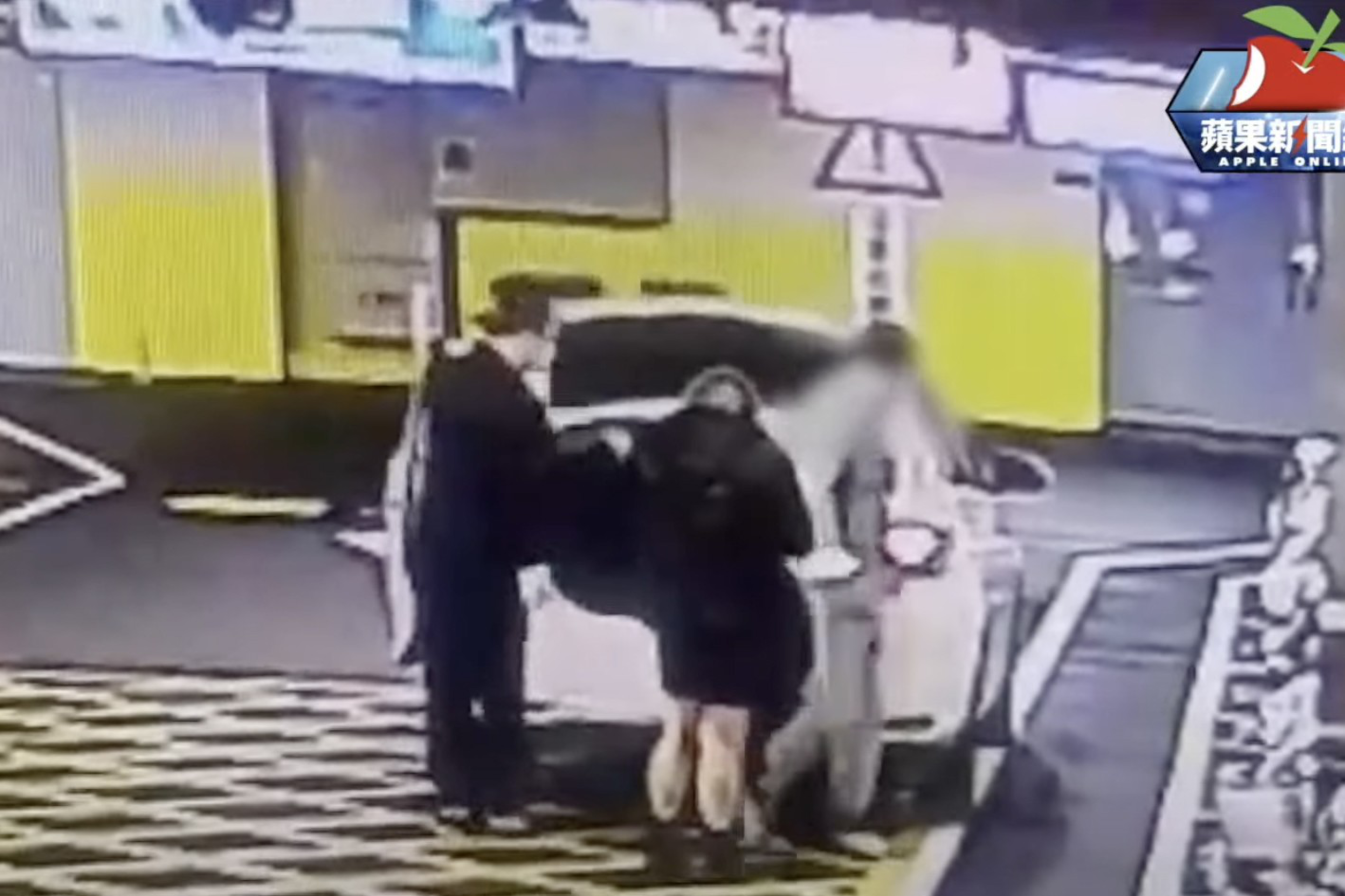 Three people stand by a white car parked near yellow buildings, with one person wearing black and another blurred in motion.