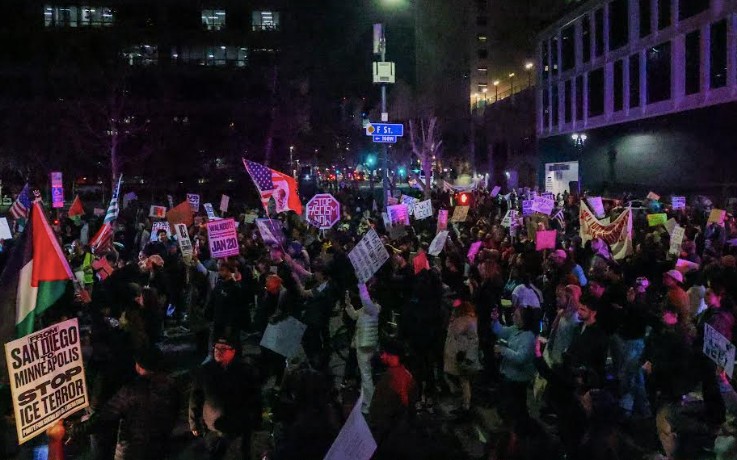 Dozens of people gather beneath a street sign holding U.S. flags and signings protesting ICE actions in Minnesota.