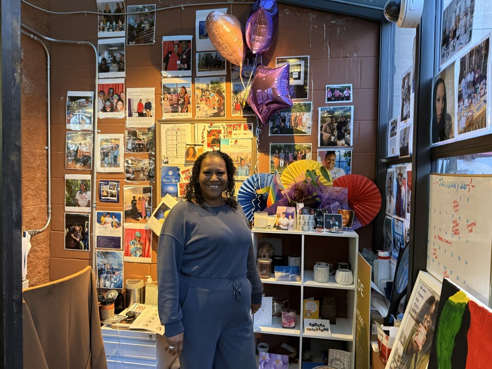 A woman wearing a blue-grey sweatshirt stand in front of a wall filled with pictures, candles and other colorful items. 
