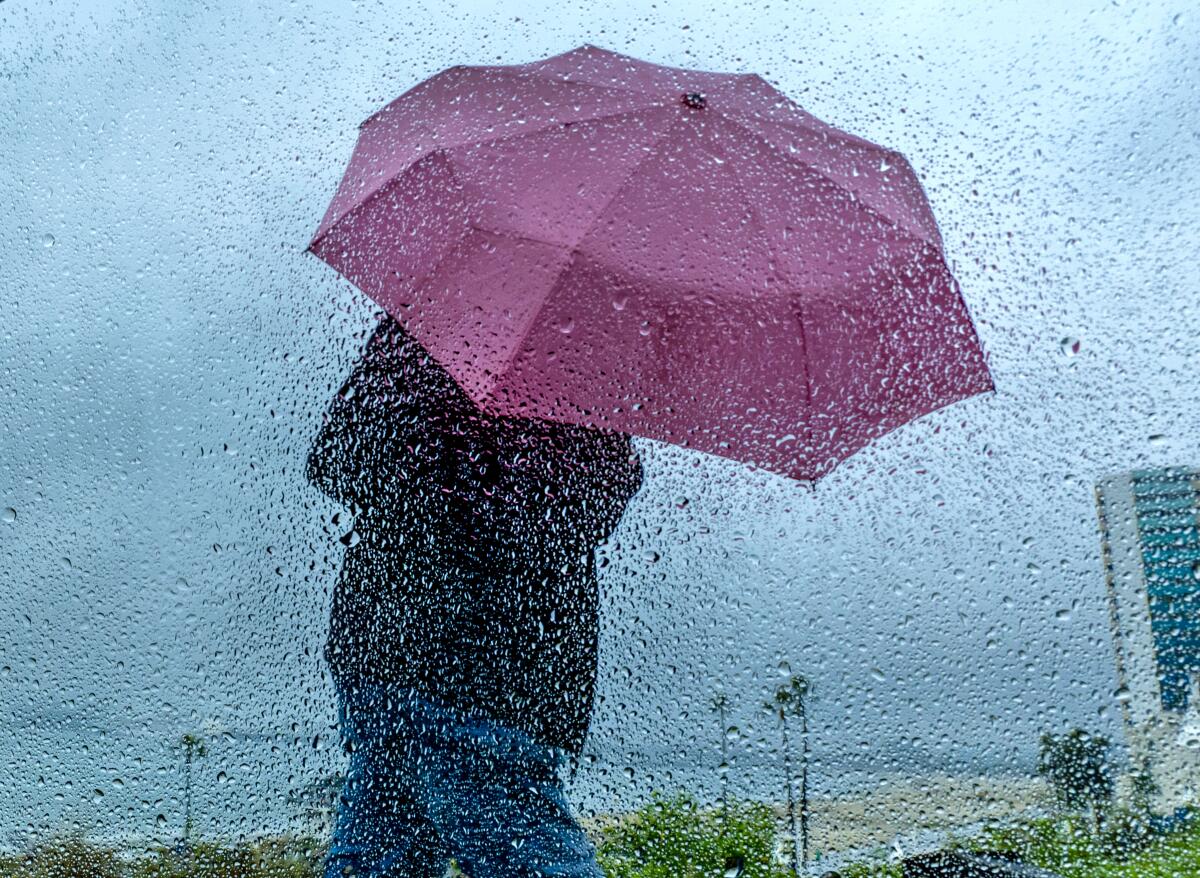 A pedestrian in a raincoat carrying an umbrella is seen through a glass window covered with raindrops.