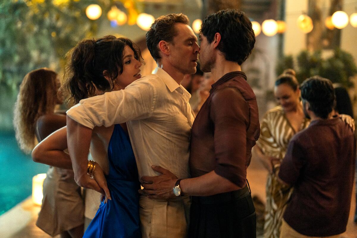 A woman in a blue dress presses against the back of a man in white, who is being held at the hips by a man in a mesh shirt.