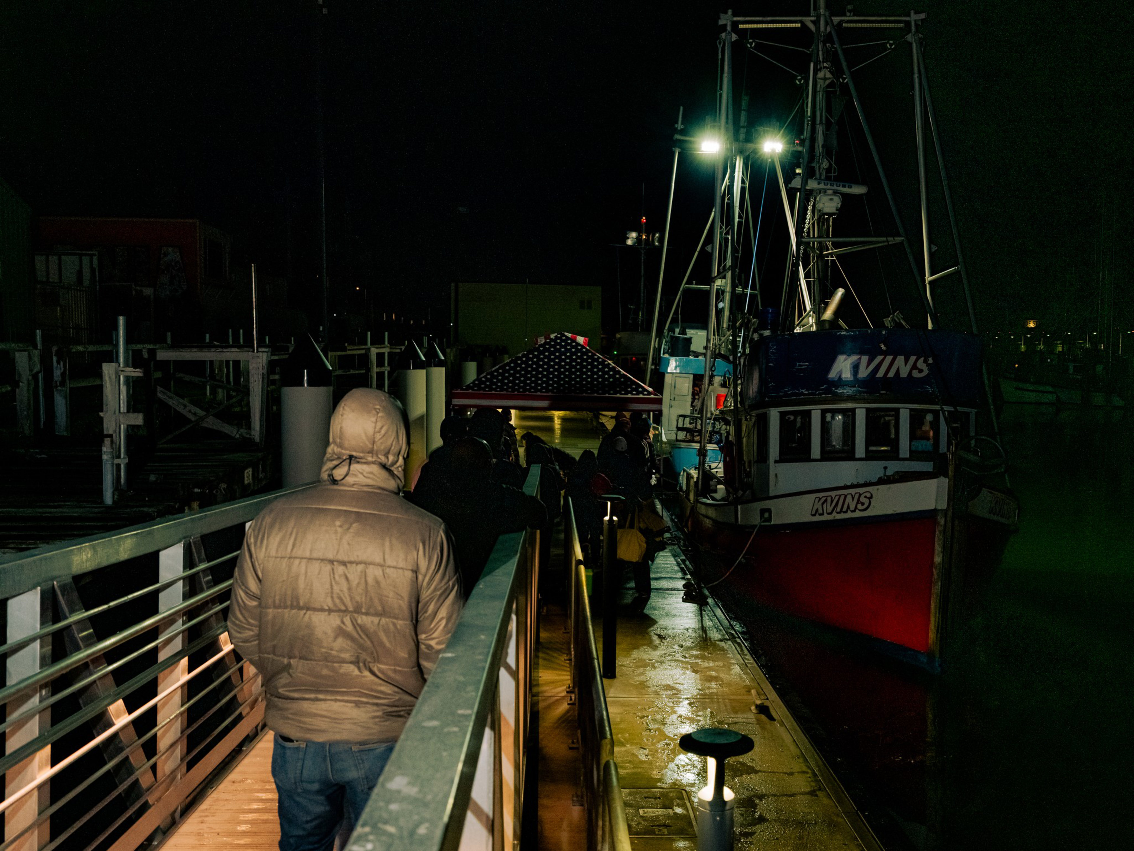 A person in a tan jacket stands on a dock at night near a red and white boat named “KVINS,” with other people and a tent in the background.
