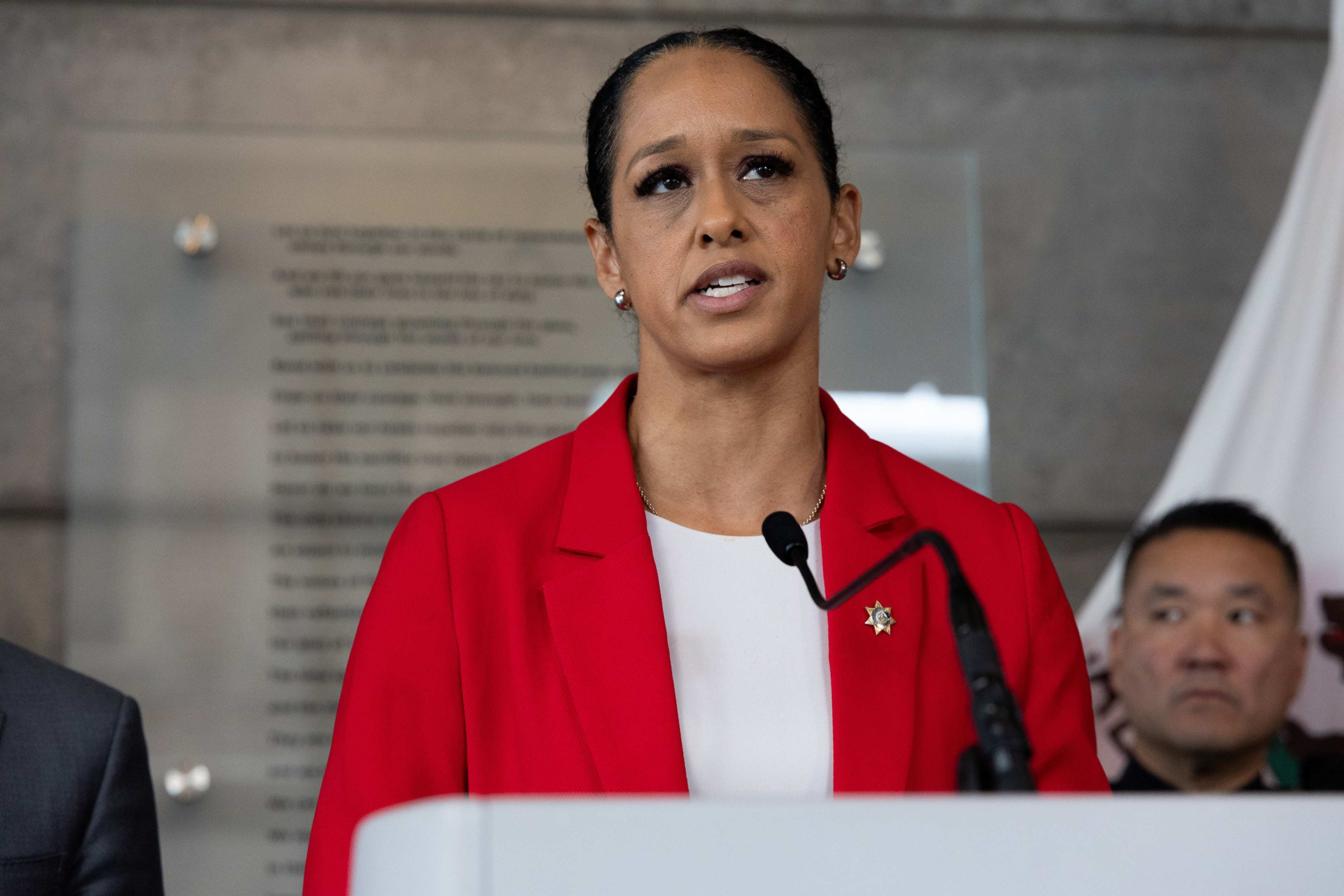 A woman in a red blazer speaks at a podium with a microphone, with a man and a blurred plaque in the background.