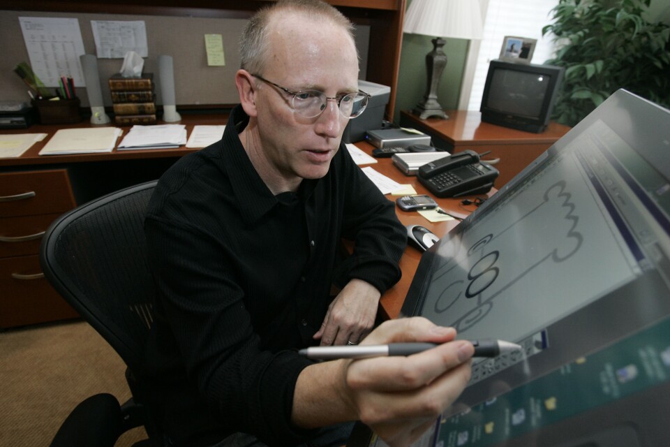 A man wearing a black shirt and eyeglasses sits at a large monitor, holding a stylus.