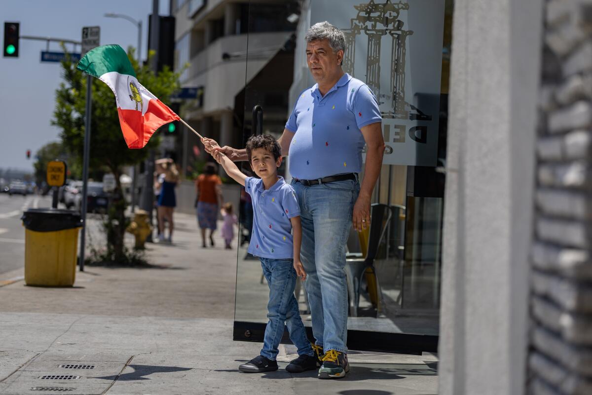 A man and a boy holding a flag stand on a sidewalk on a sunny day
