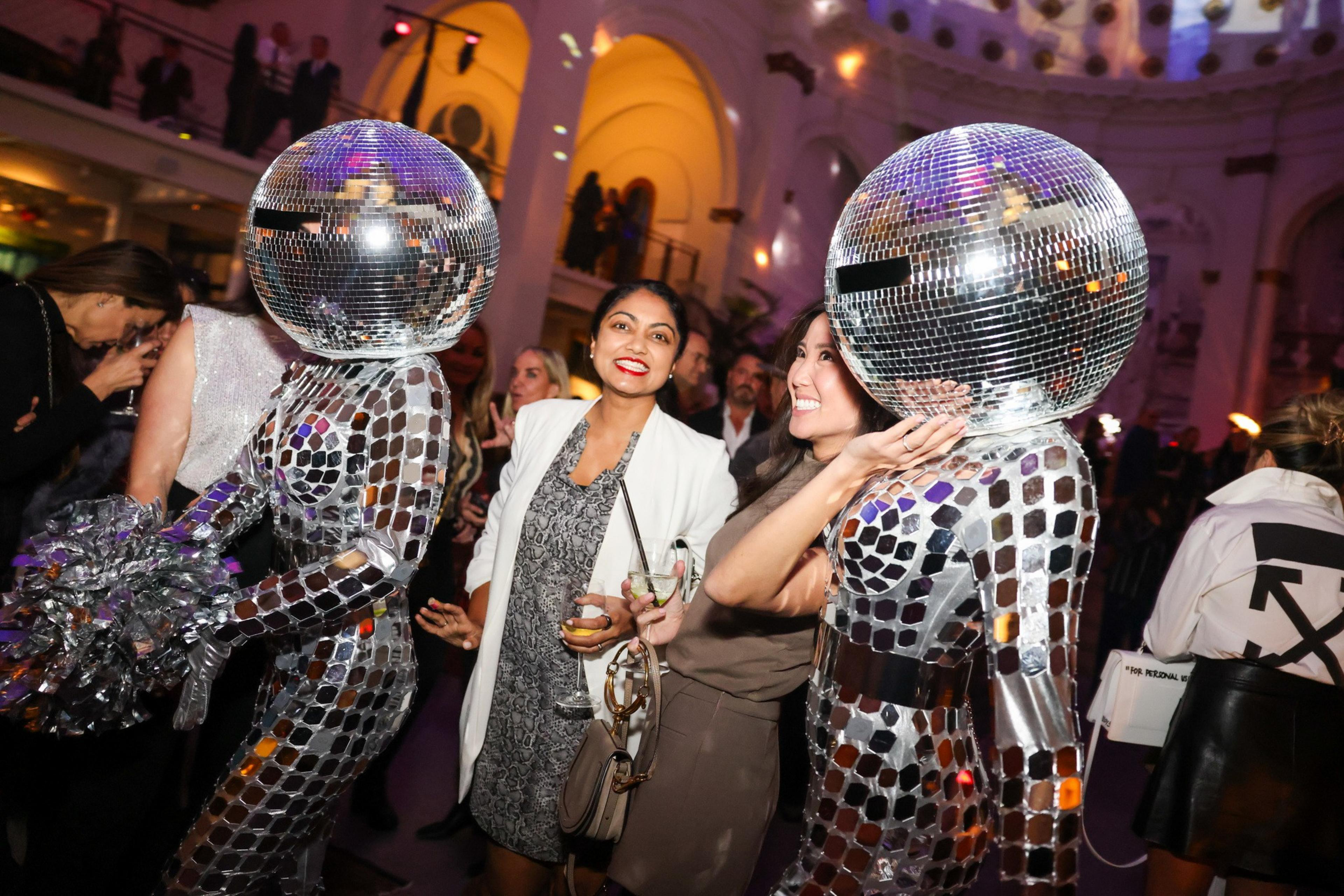 People in a lively venue with two individuals wearing mirror-tiled outfits and disco ball helmets. Others are dressed in formal attire, enjoying the festive atmosphere.