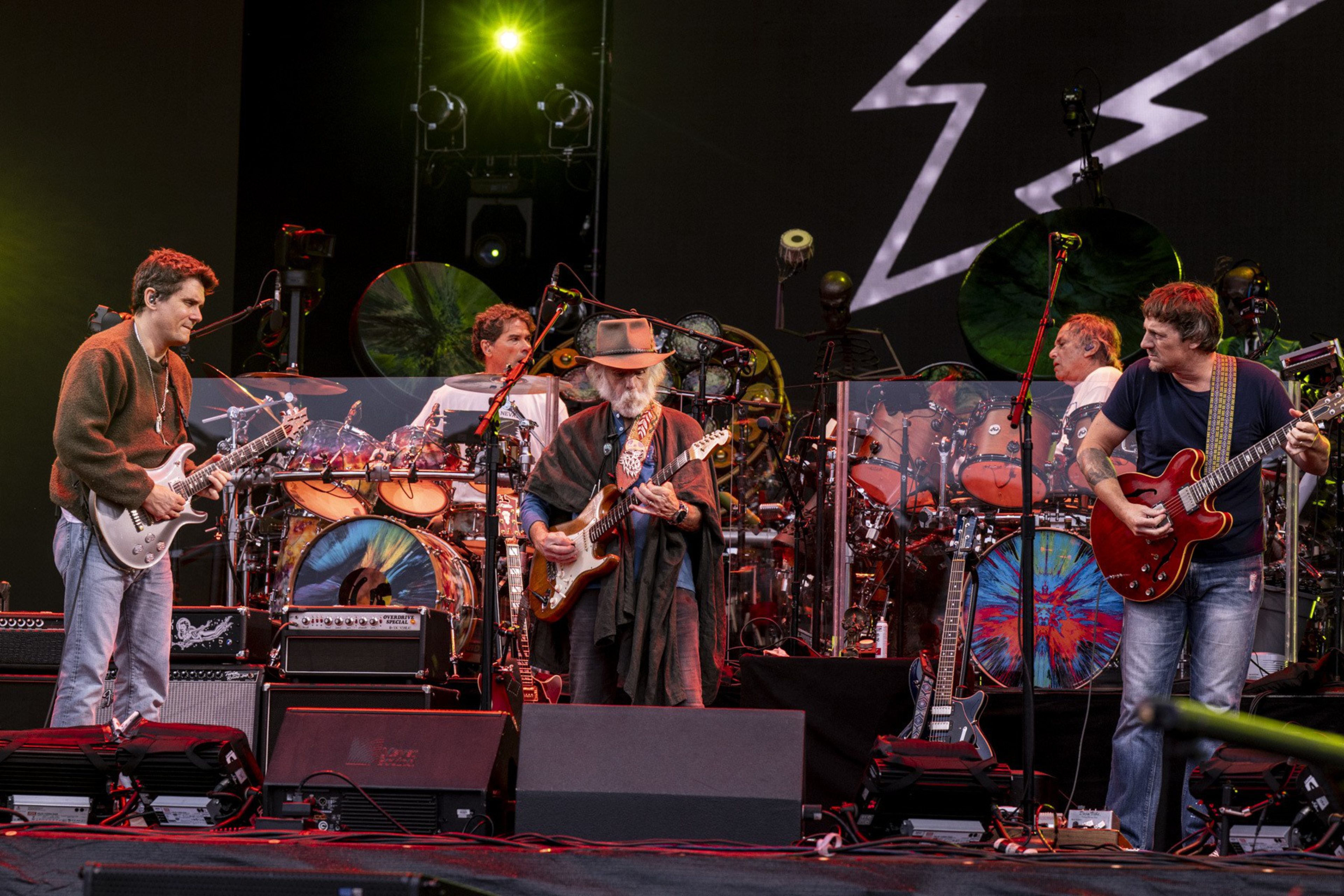 Five musicians perform on stage with guitars and drums, colorful drum heads, stage lights, and a large lightning bolt graphic in the background.