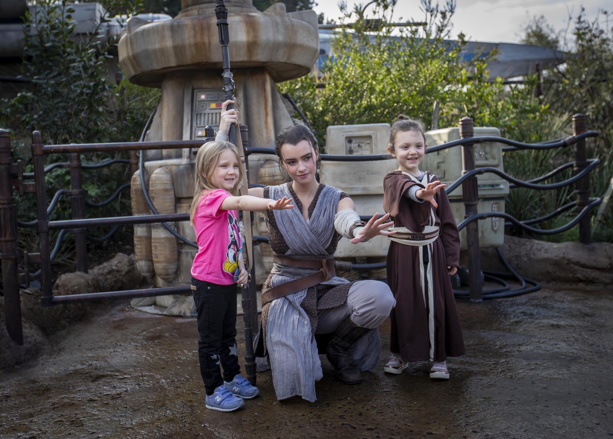 A woman dressed as Rey from "Star Wars" kneels and poses with her hand outstretched for a photo with two little girls.