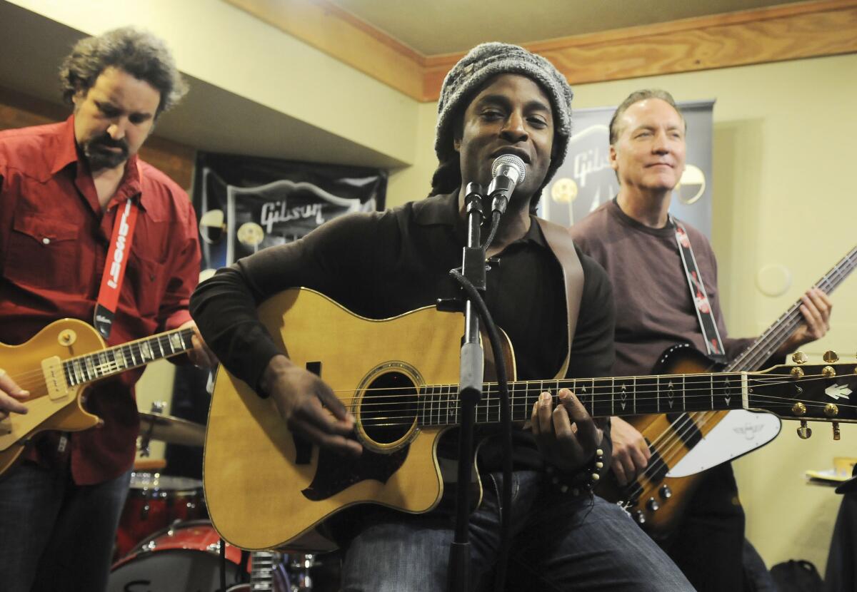 A man sitting and playing guitar with two other musicians in the background.