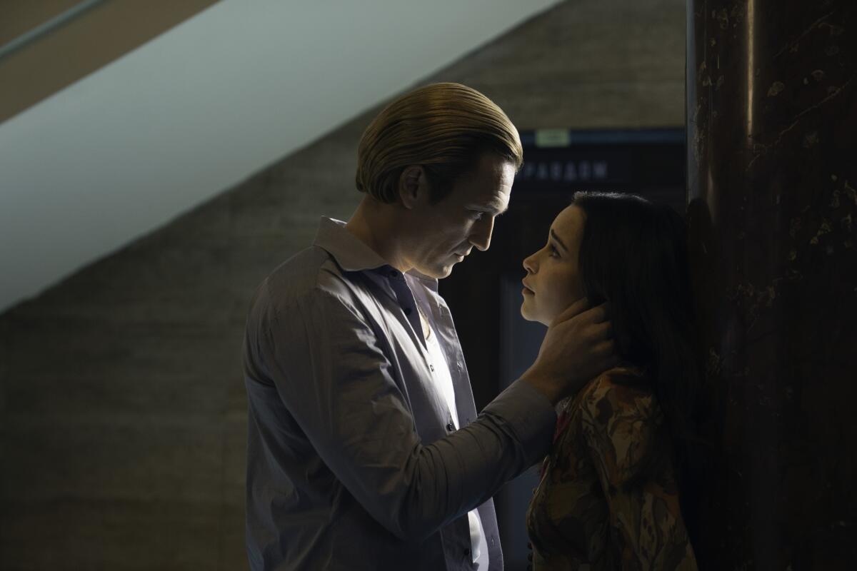 A man holds a his hand near a woman's neck, who is standing against a wall.