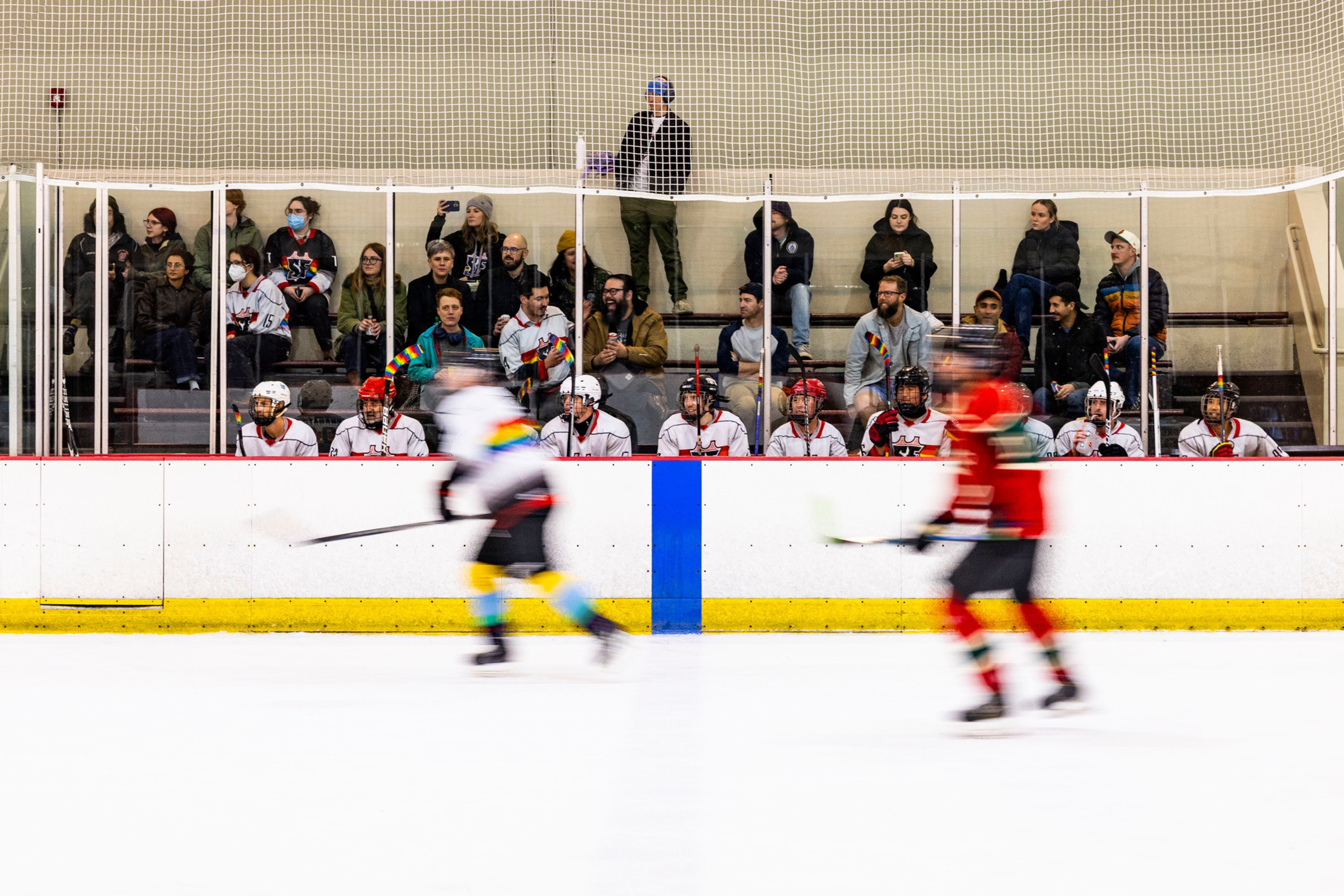 Hockey players wearing white and red uniforms sit on the bench while two blurred players skate on the ice in front, with spectators watching behind protective glass.