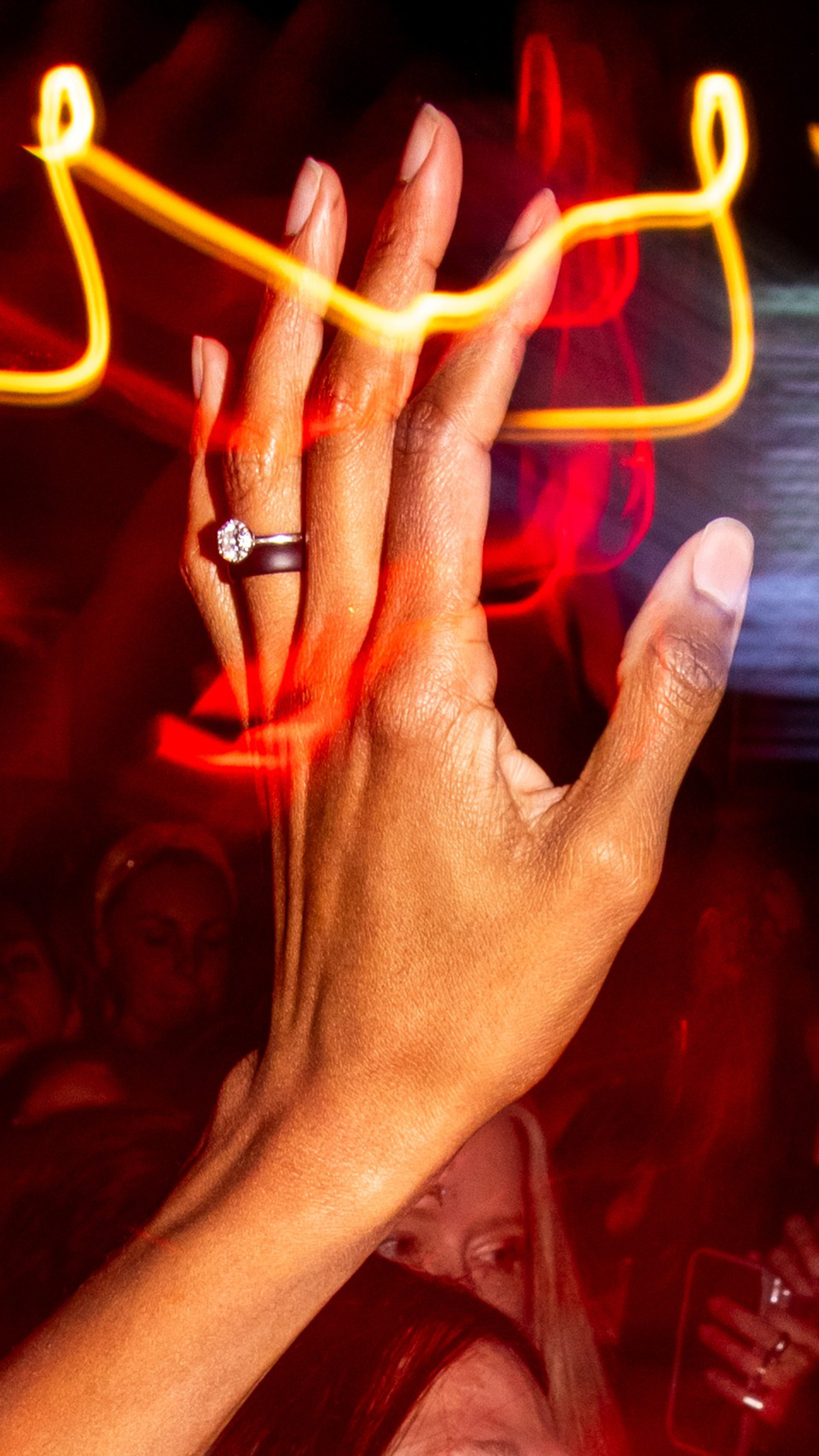 A raised hand with a diamond ring on the ring finger is highlighted by streaks of orange and red light against a dark background.