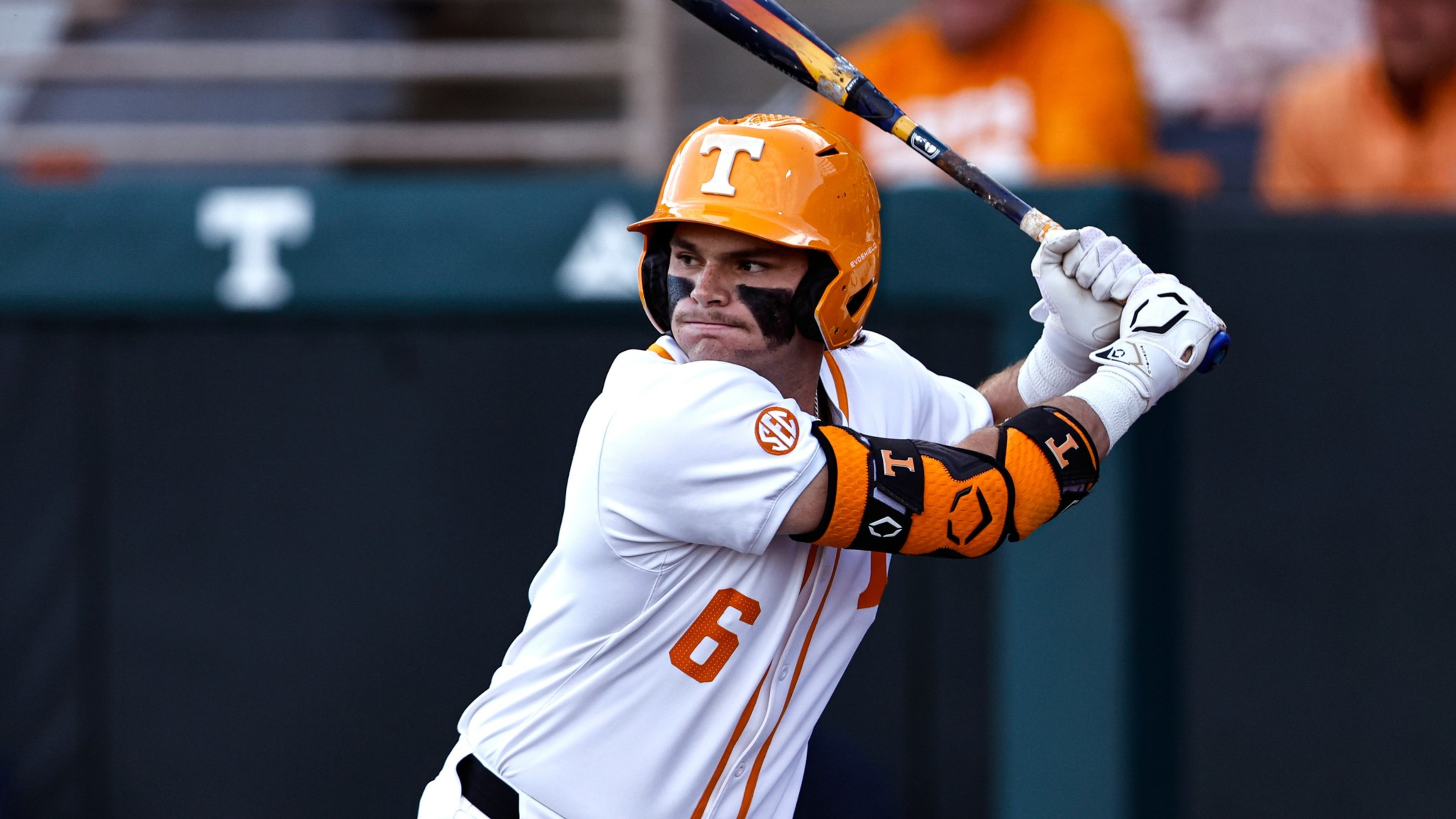 A baseball player in a white uniform and orange helmet is poised to bat. He wears eye black, gloves, and an arm guard, against a blurred stadium backdrop.