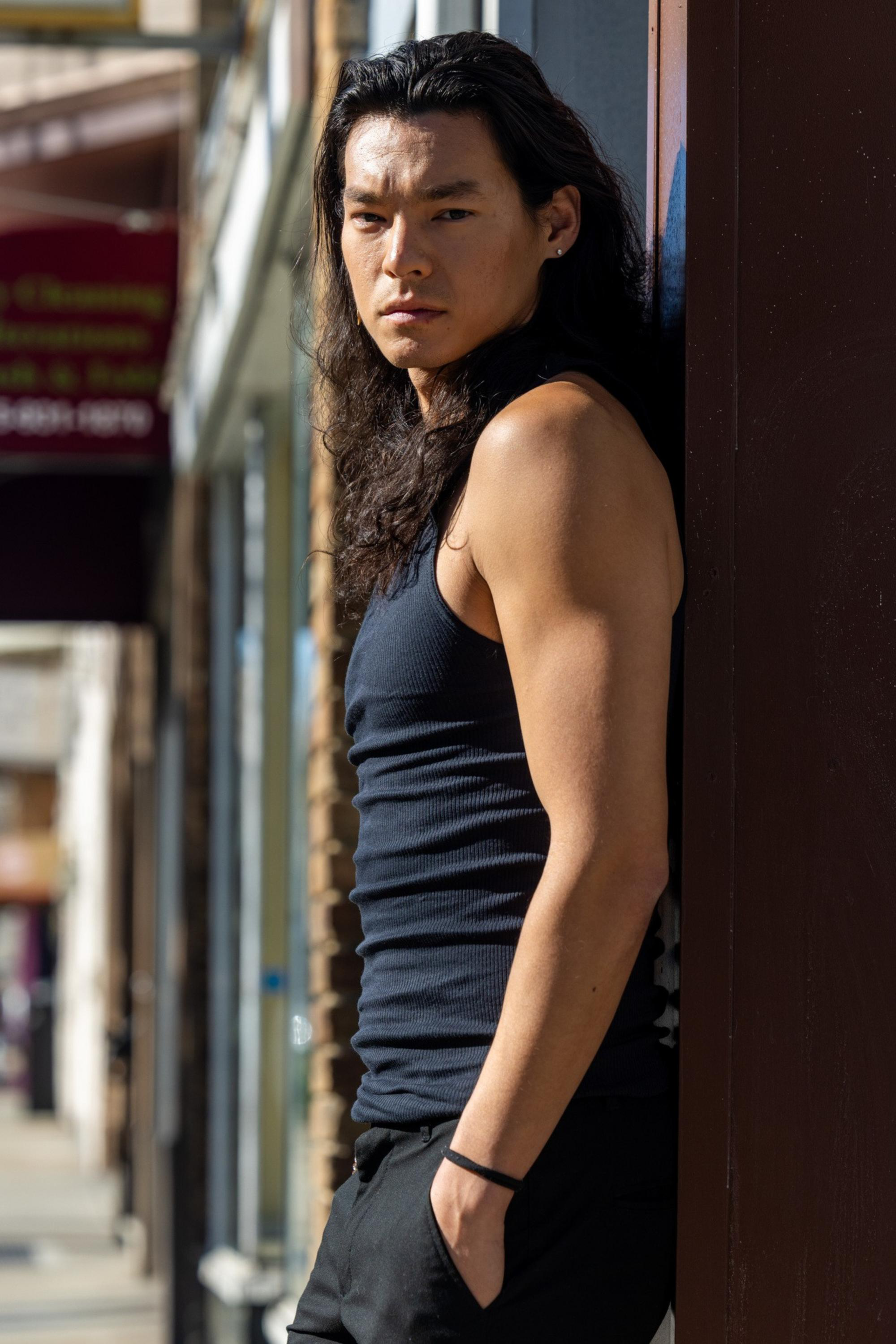 A young man with long dark hair leans against a wall outdoors, wearing a black tank top and black pants, looking intently at the camera.