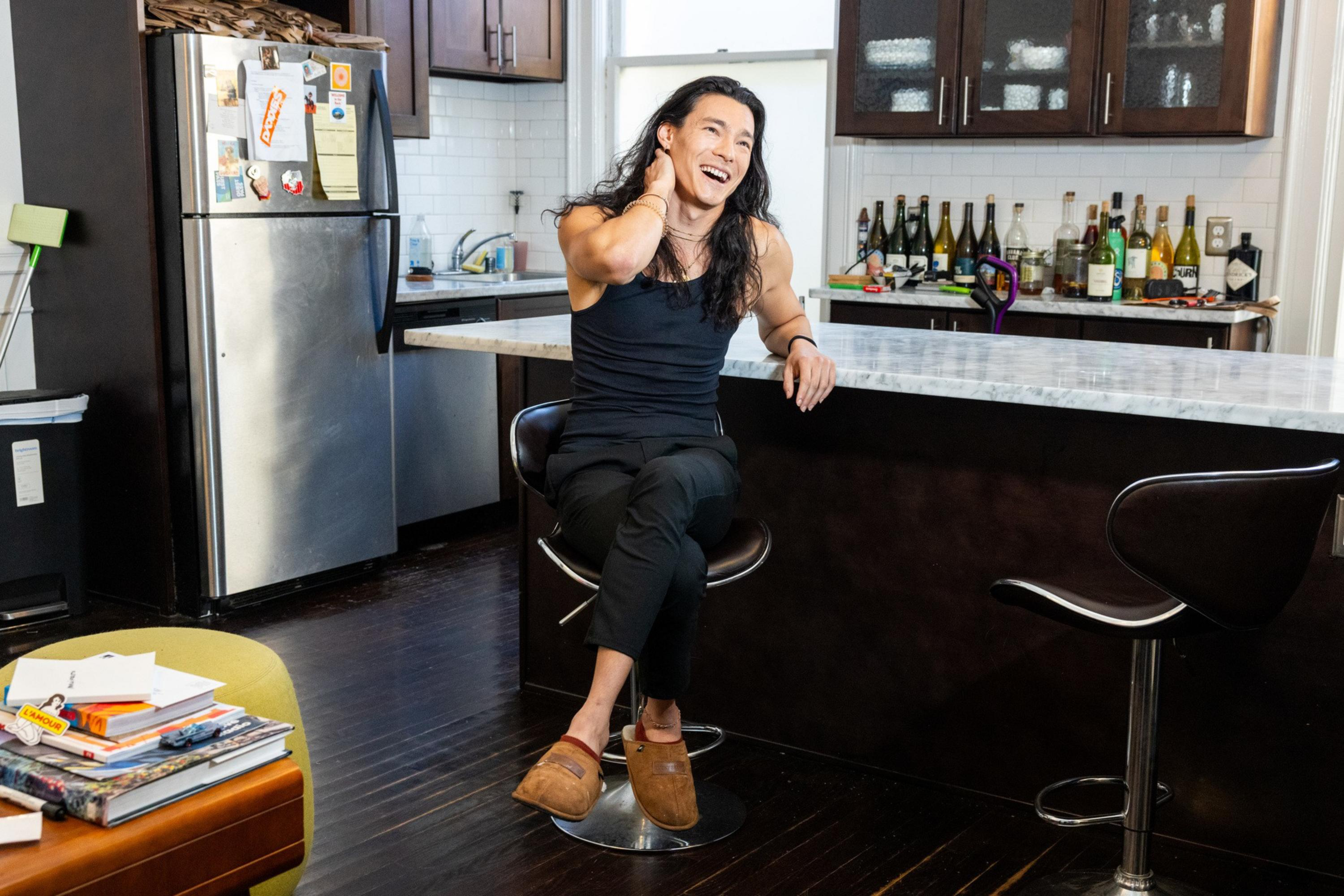 A person with long hair wearing a black tank top and pants sits on a bar stool in a kitchen, laughing with one hand on their neck.