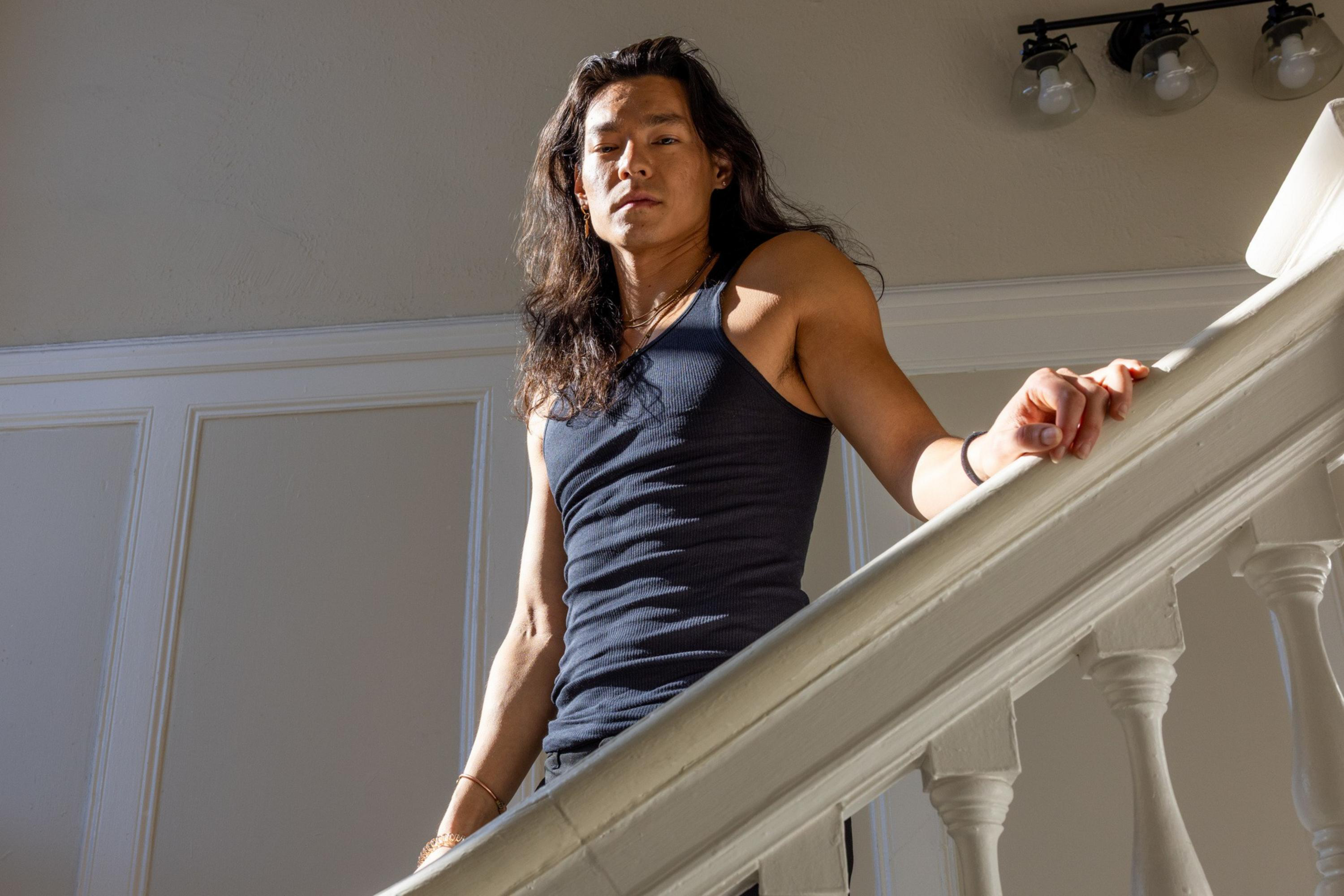 A person with long dark hair wearing a black sleeveless shirt leans on a white banister, looking down with sunlight casting shadows.