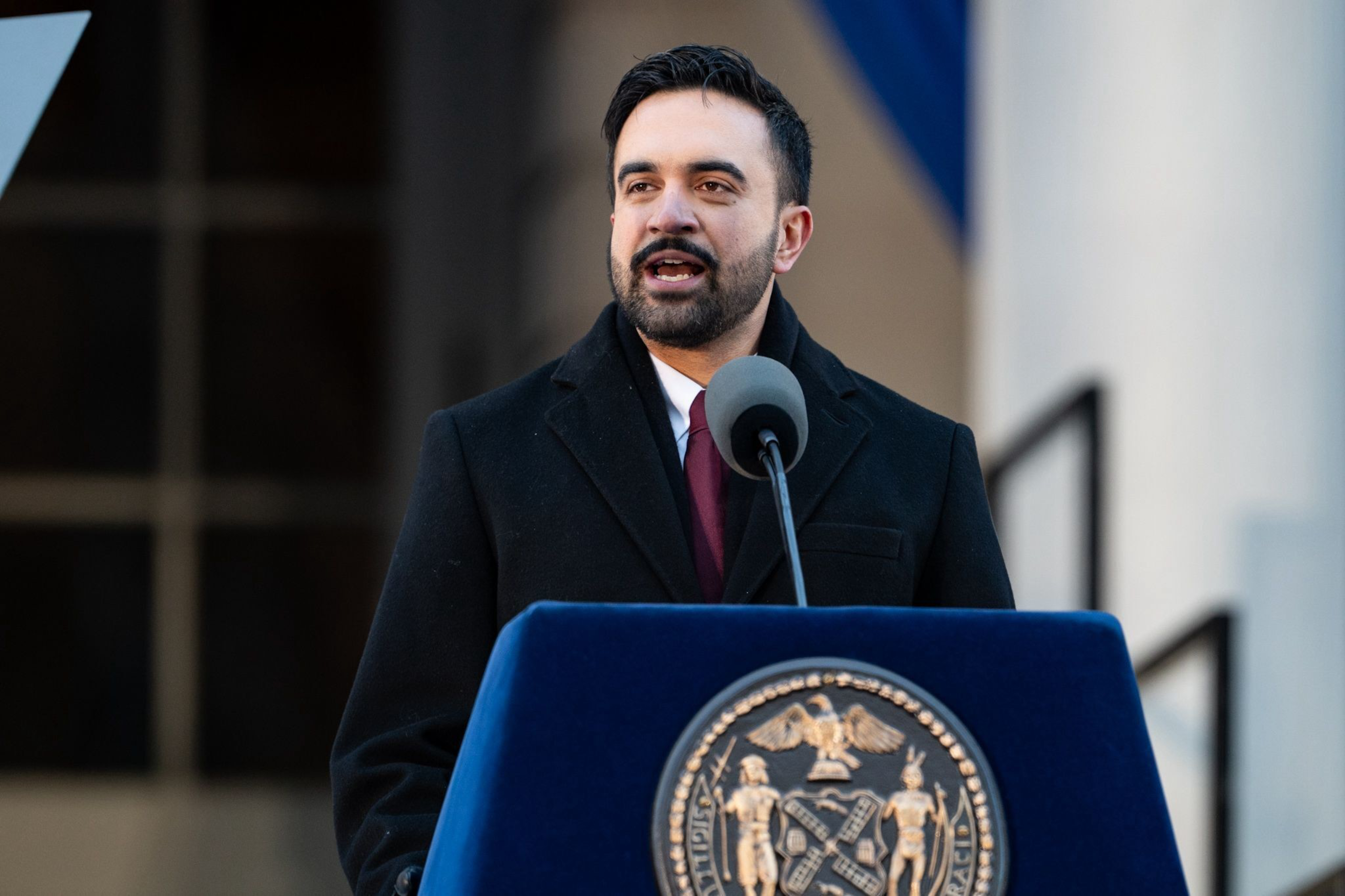 A man with dark hair and a beard, wearing a black coat and burgundy tie, speaks at a podium with an official seal in front of a building.