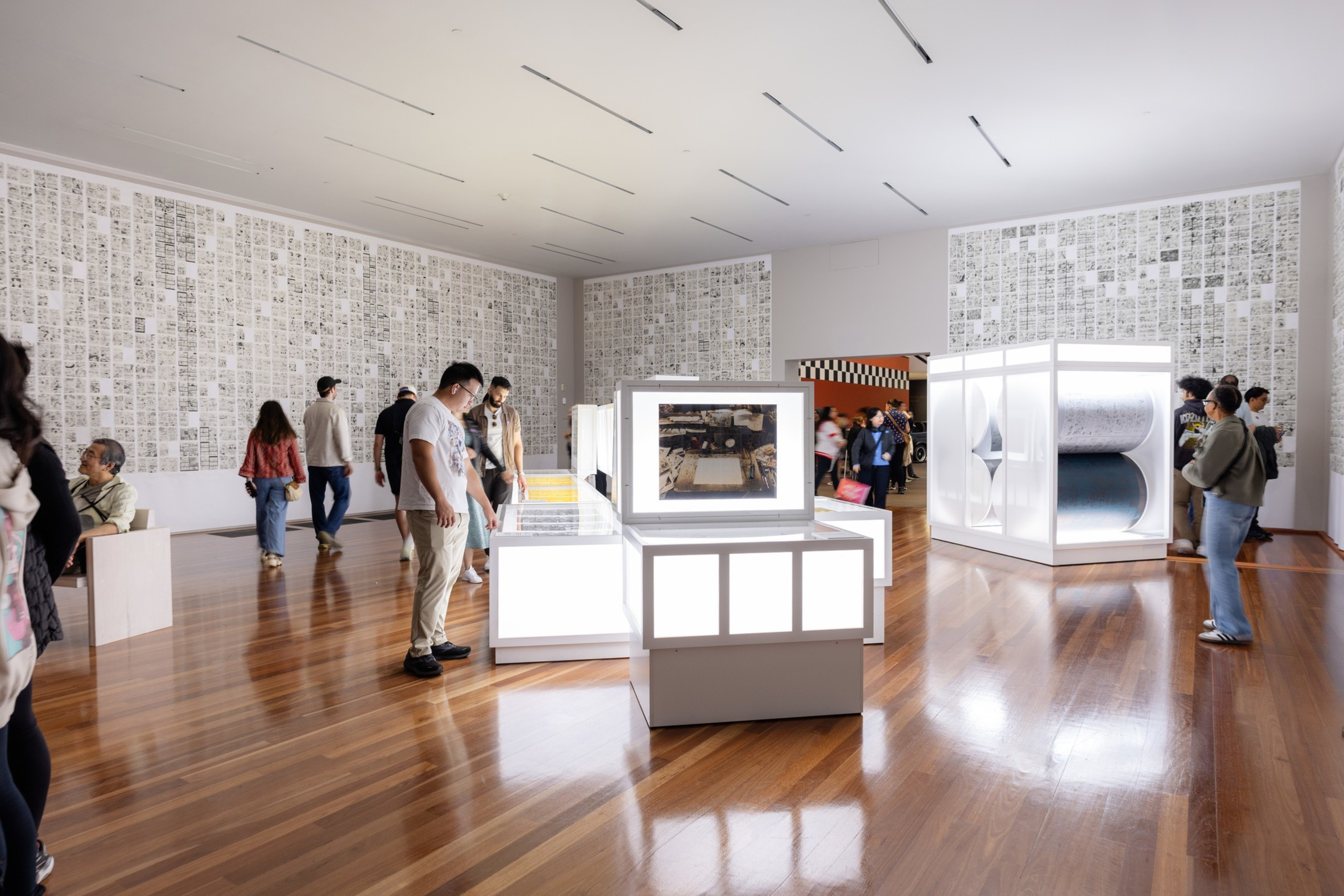 People explore a well-lit gallery with large illuminated display cases and walls covered in grids of small, detailed black-and-white drawings or prints.